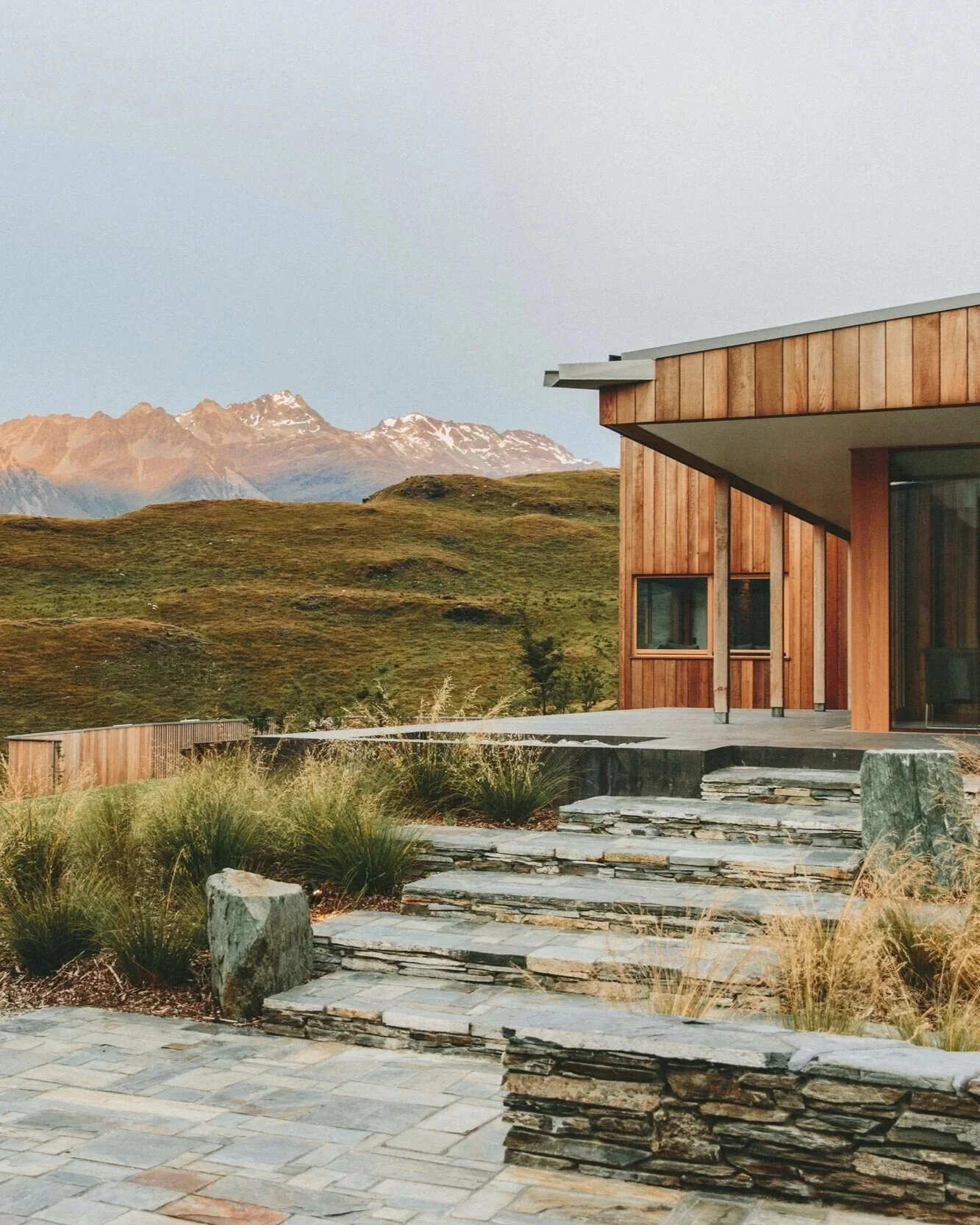 Modern wooden house with stone steps leading to a patio, set in a rural landscape with rolling hills and snow-capped mountains in the background.