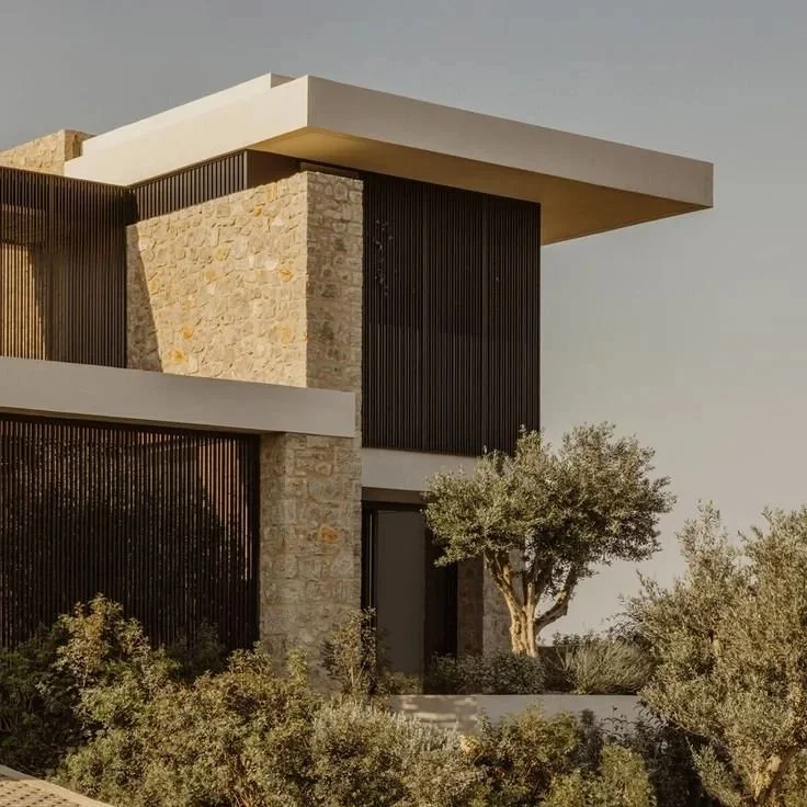 Modern house with stone, white, and black elements, surrounded by trees and bushes.