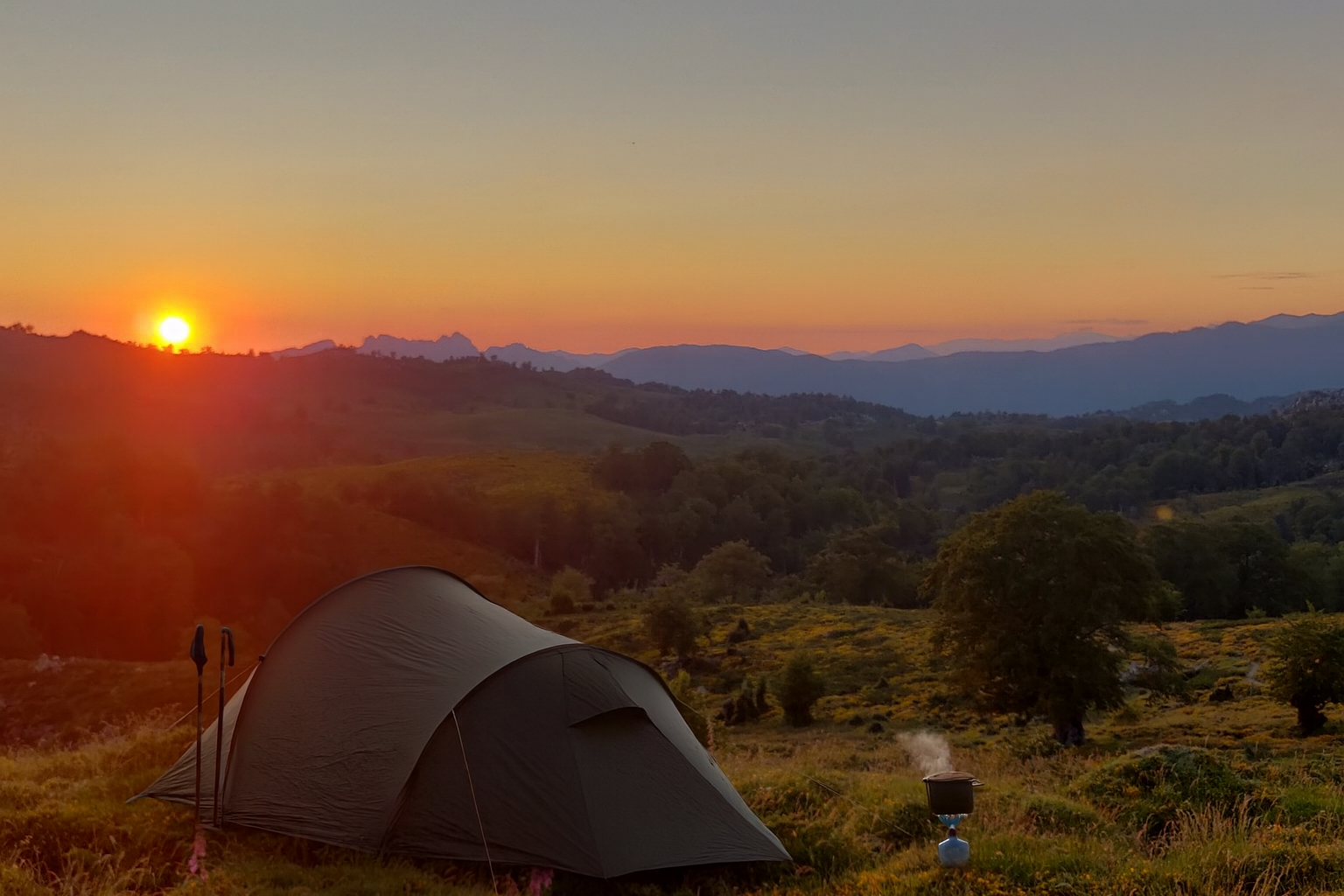 Tente de camping sur une colline verdoyante au coucher du soleil, avec un paysage de montagnes en arrière-plan et un petit poêle à bois fumant à proximité.