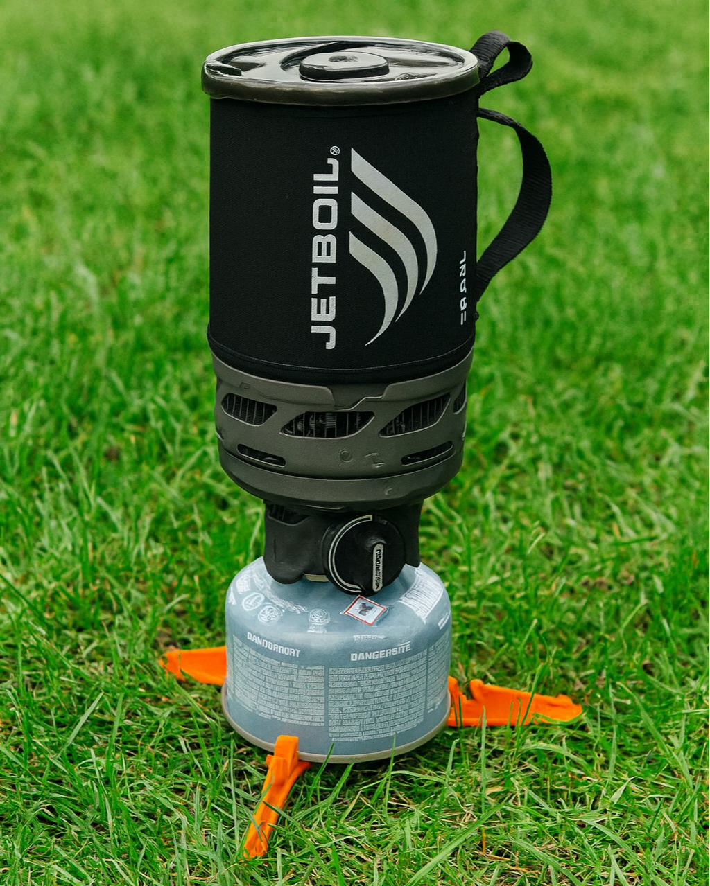 Un réchaud à gaz avec une tasse suspendue dessus, placé sur de l'herbe verte, utilisé pour la cuisine en plein air.