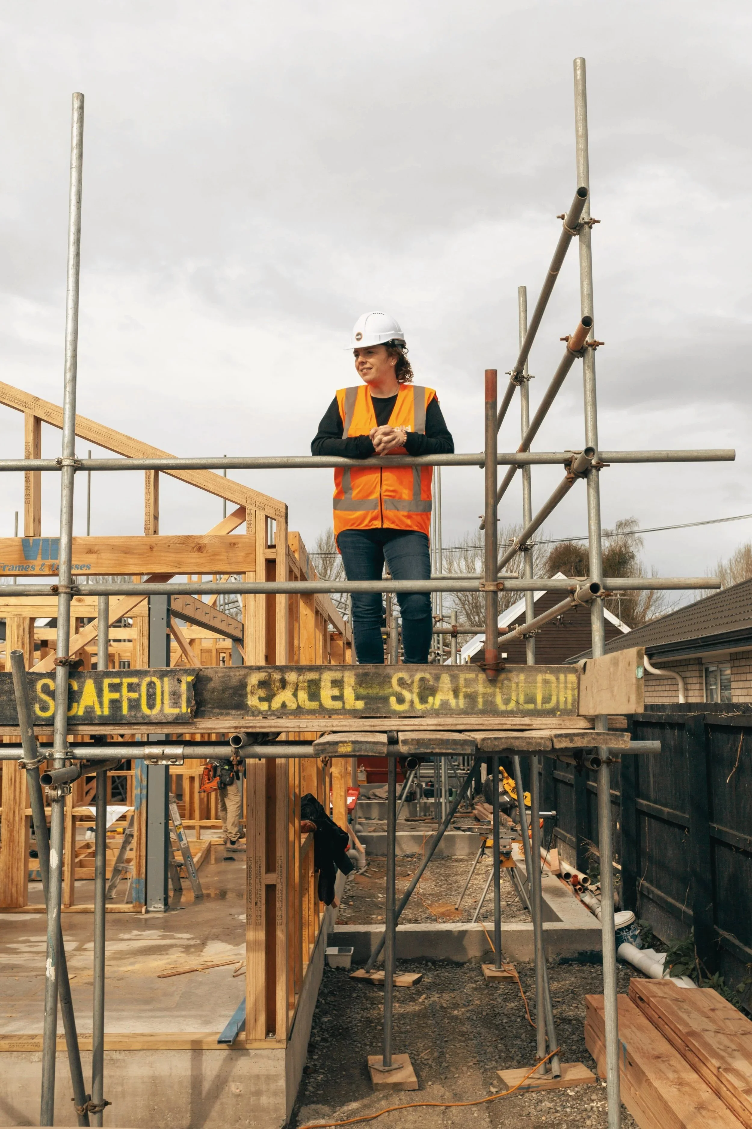 A woman wearing a white safety helmet and orange safety vest stands on a building construction site, overlooking the framework of a wooden structure with scaffolding around her, under a cloudy sky.