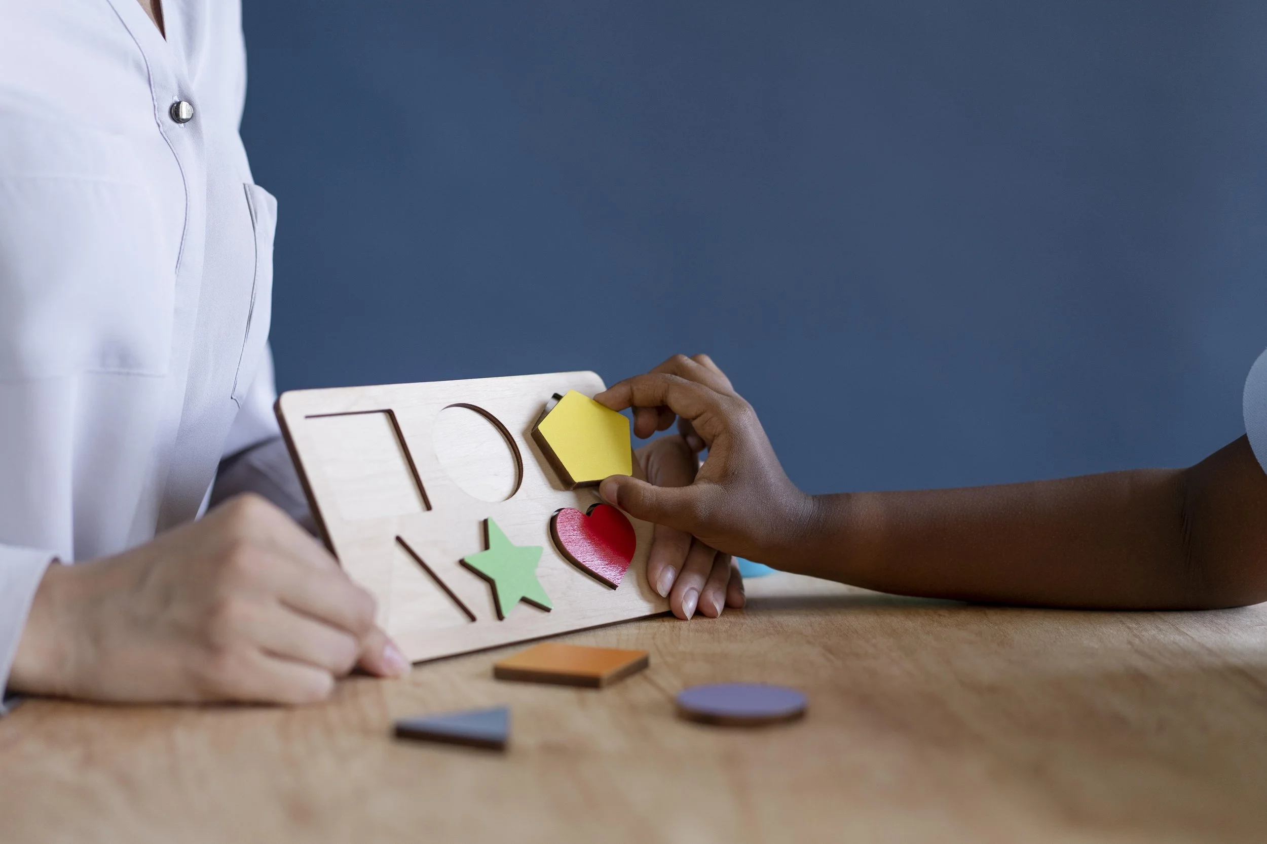 A person offering a child's hand a wooden shape puzzle with colorful pieces, including a yellow hexagon, a red heart, a green star, and other shapes, on a wooden table.