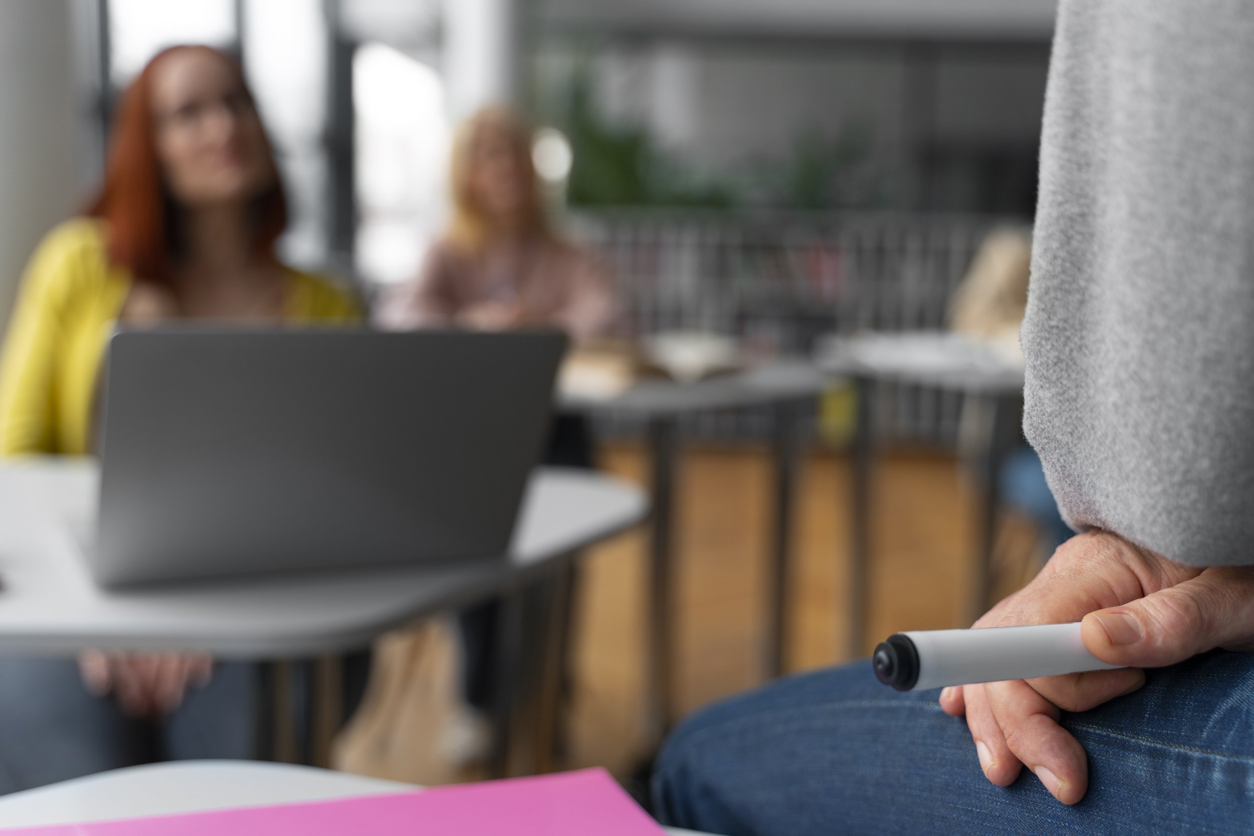 Person holding a marker in their hand, sitting near a table with a pink paper, with two women blurred in the background, one wearing a yellow top and the other in pink, in a room with large windows and a metal railing.