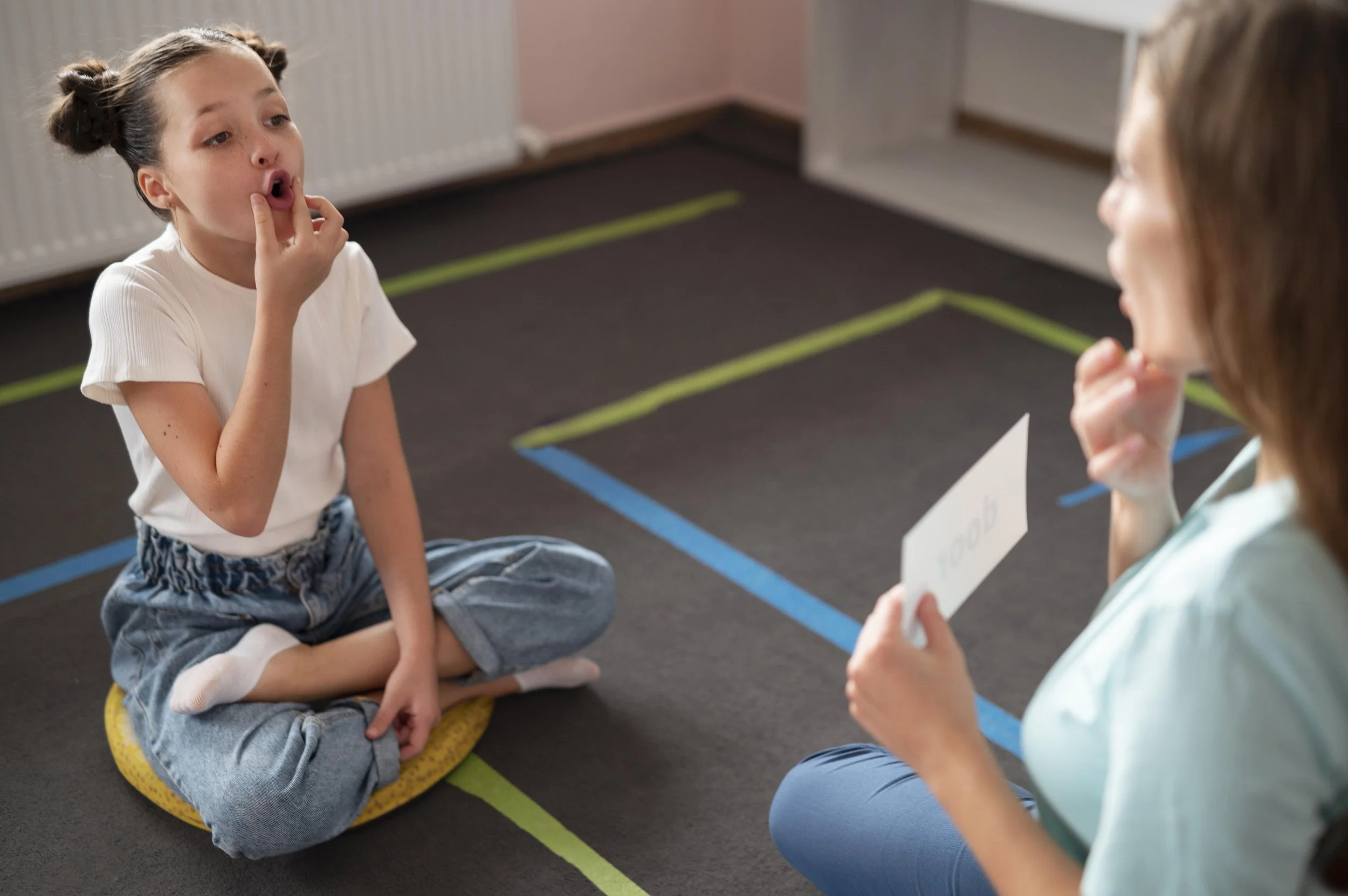 A young girl sitting cross-legged on a yellow cushion on the floor, touching her face, listening to a woman holding a card, in a room with colorful tape markings on the carpet.