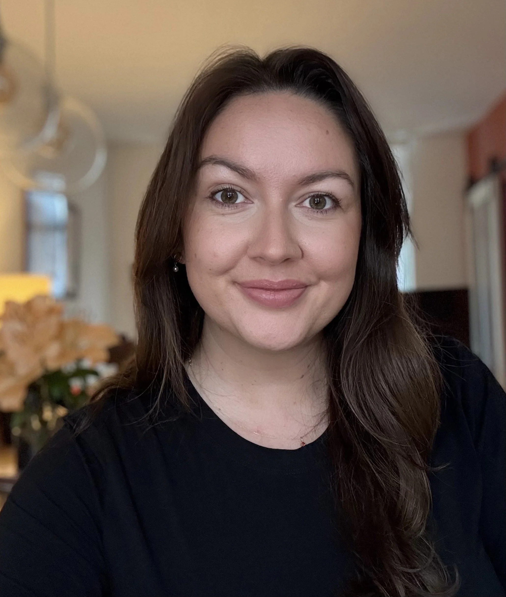 A woman with long brown hair and brown eyes, smiling, wearing a black top, indoors with blurred background of a living room.