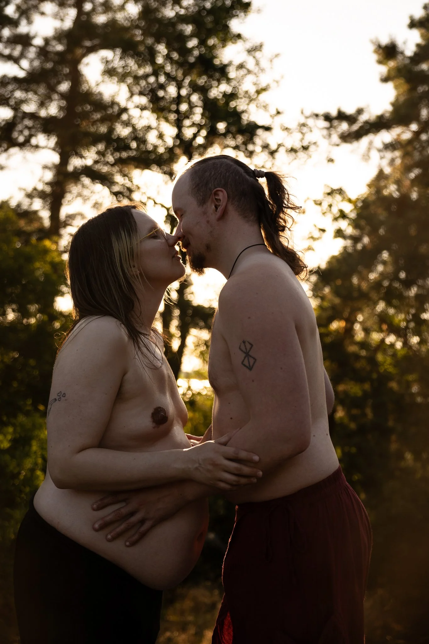 Kaksi isää, joista toinen on raskaana, kasvotusten lähellä suudelmaa. Kuva on lämpimän sävyinen ja kevyt vastavalo