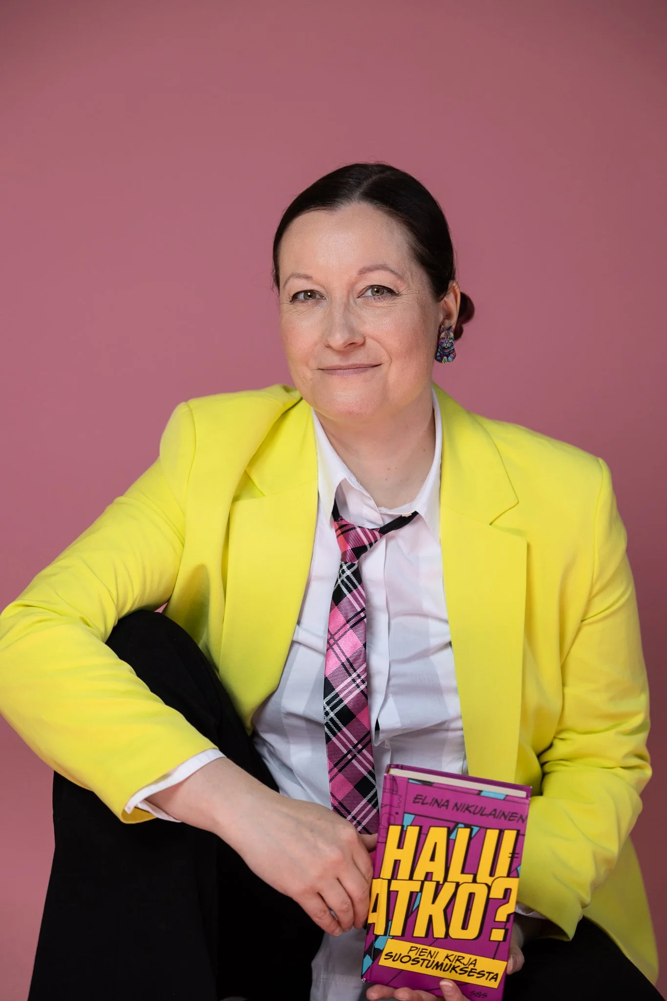 Laaja puolikuva, valkoinen henkilö istuu toinen jalka tuolille nostettuna ja pitää kädessään kirjaa "Haluatko? Pieni kirja suostumuksesta". Tausta on vaaleanpunainen, kuvattavalla on päällään keltainen jakku, valkoinen kauluspaita ja pinkki kravatti.