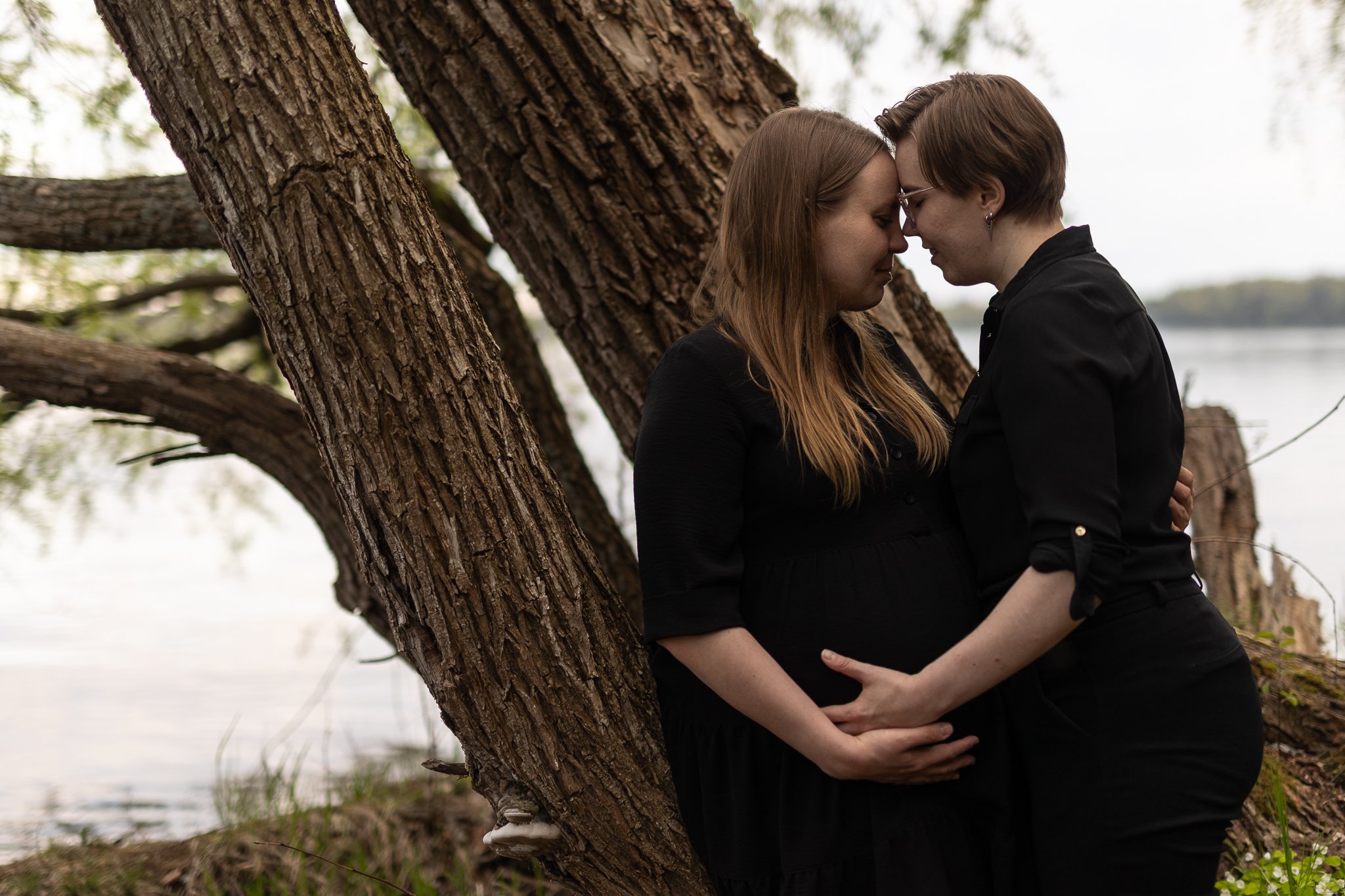 Odotuskuva järven rannalla, odottaja nojaa puuhun, kumppani kasvotusten, otsat yhdessä