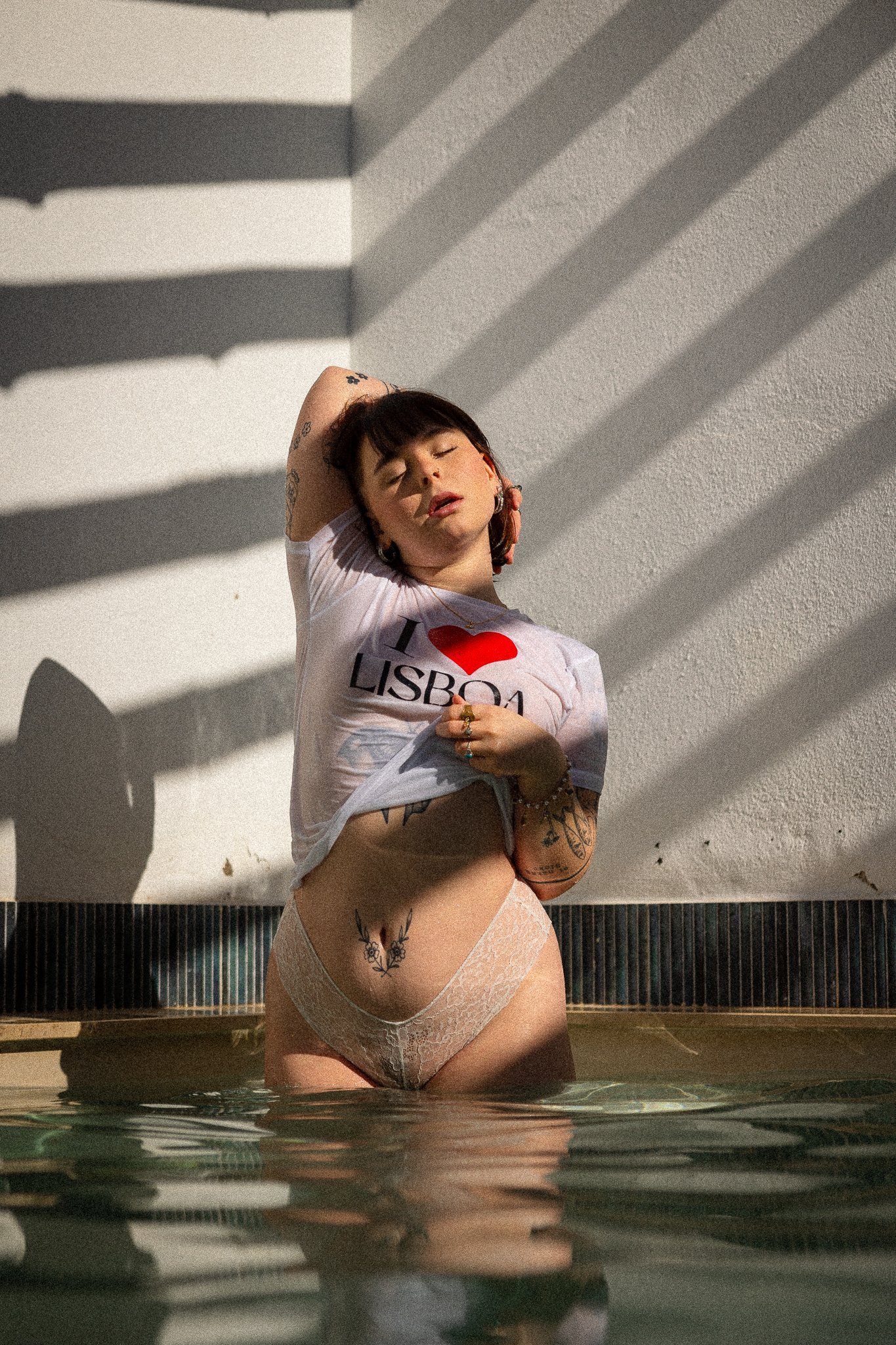 Henkilö seisoo silmät kiinni uima-altaassa reisiään myöten, ja nostaa toisella kädellä vatsaltaan paitaa, jossa lukee I <3 Lisboa. Toinen käsi on niskassa. Hänellä on paljon koruja ja tatuointeja, aurinko paistaa "katto"paalujen välistä kirkkaana.