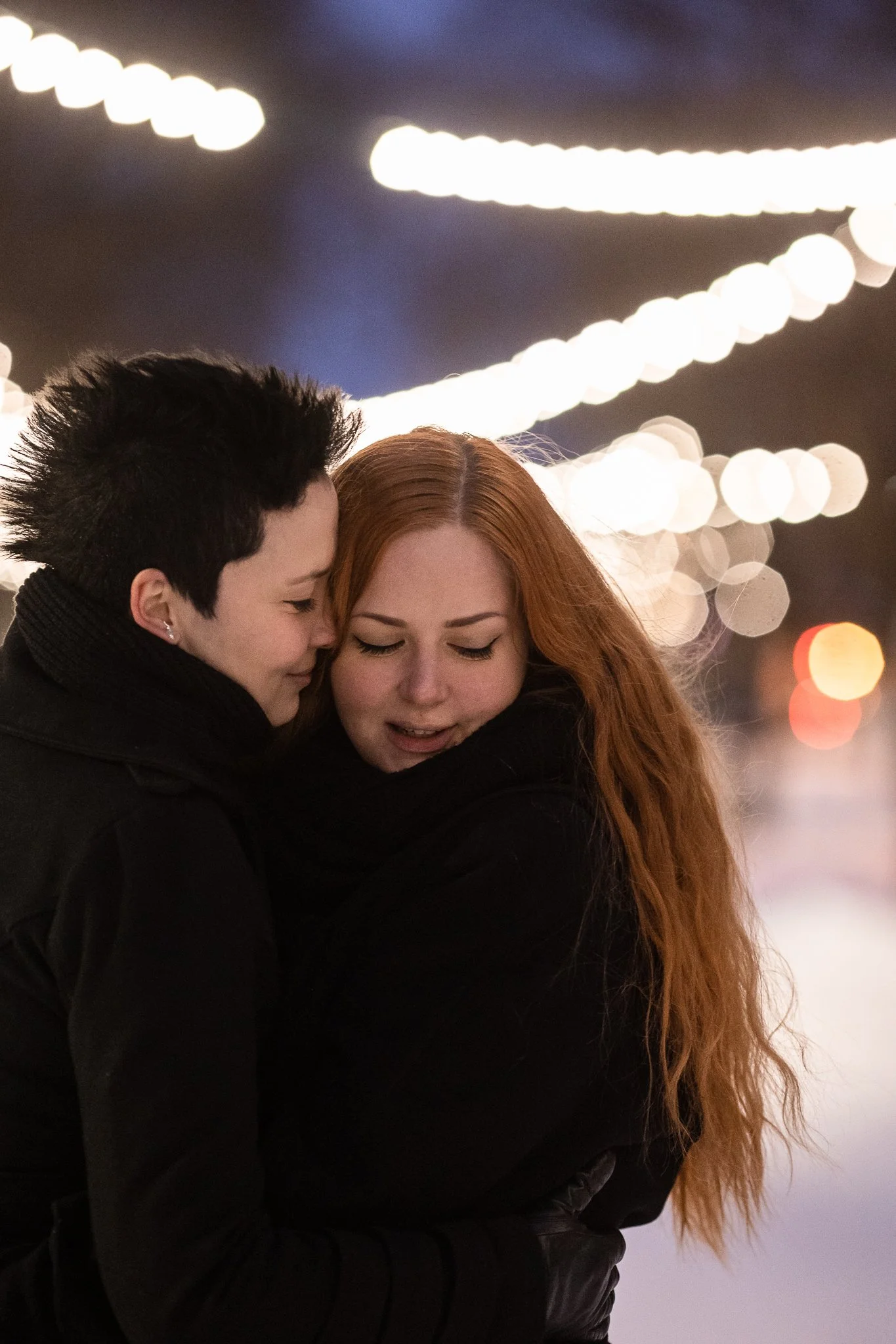 pariskunta, toisella katse alaviistoon, toinen on painautunut lähelle toisen pään sivua, taustalla näkyy valoja