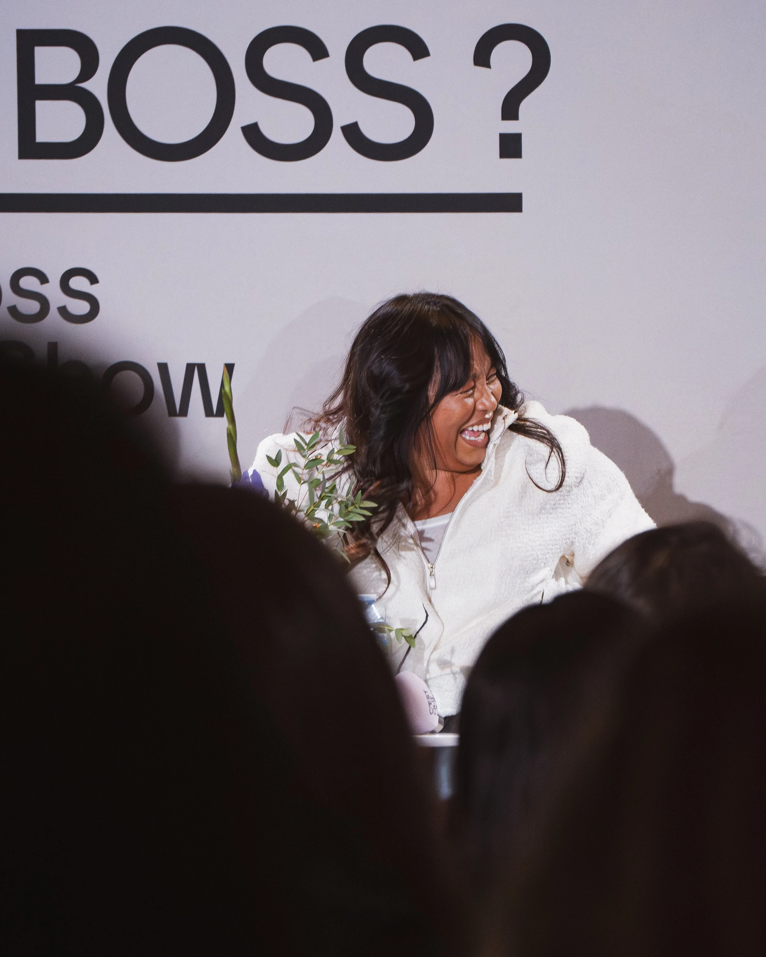Une femme souriante, vêtue d'une veste blanche, rit lors d'un événement face à un public. À l'arrière-plan, un panneau avec le mot 'BOSS ?' en gros caractères.