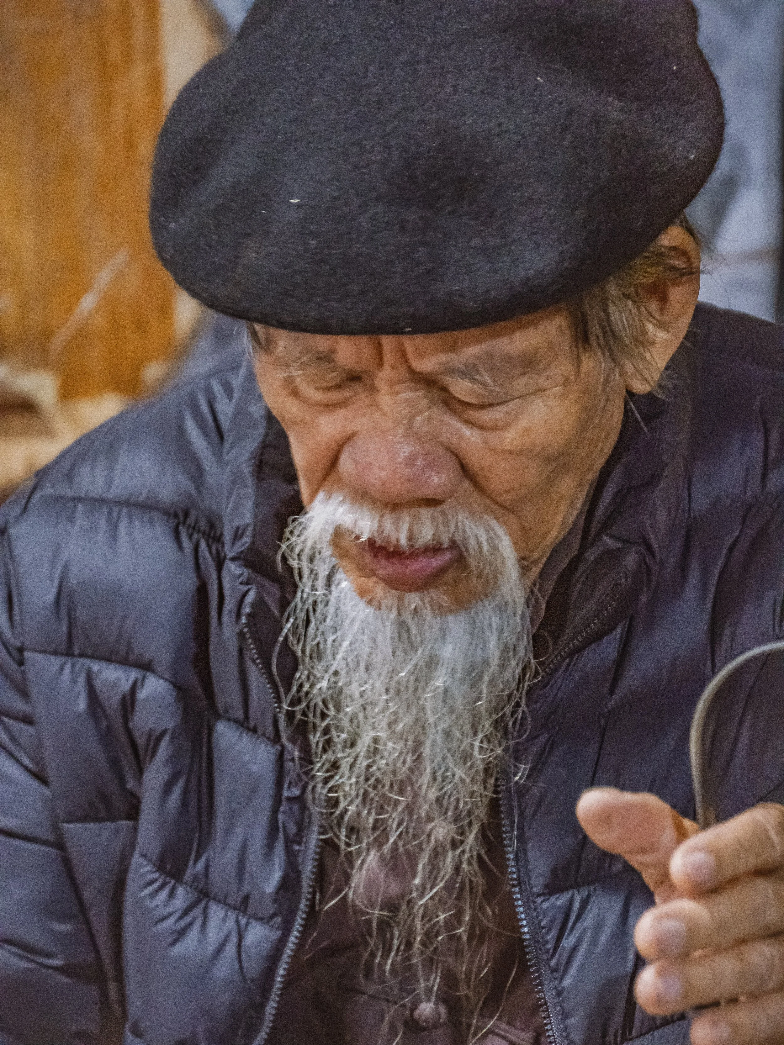 Un homme âgé portant un béret noir, une barbe blanche, une moustache et une veste noire, regardant vers le bas, en intérieur.