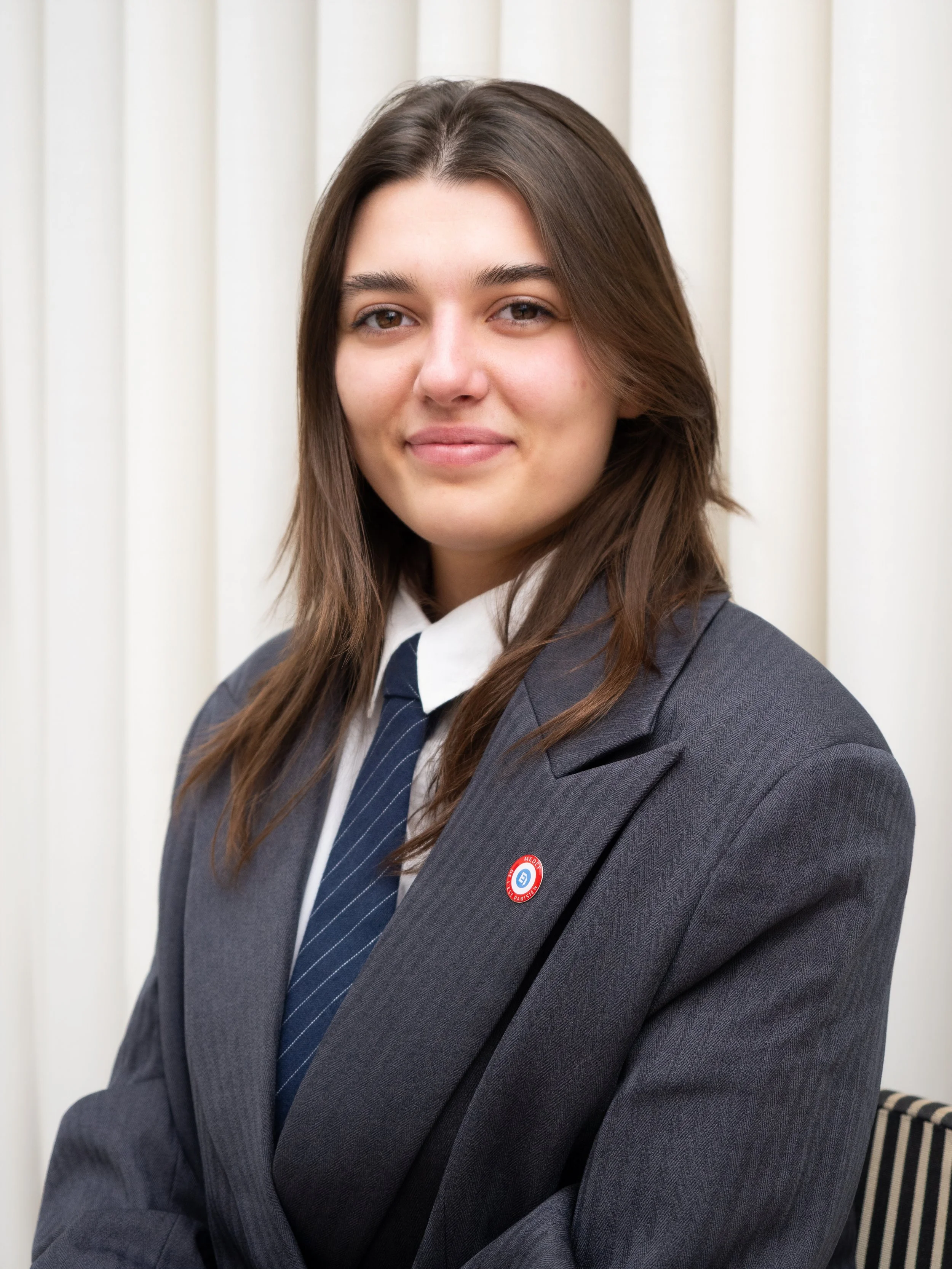 Jeune femme souriante en costume gris, chemise blanche et cravate, portant un pin avec un logo bleu et rouge, posant contre un fond de rideaux blancs.