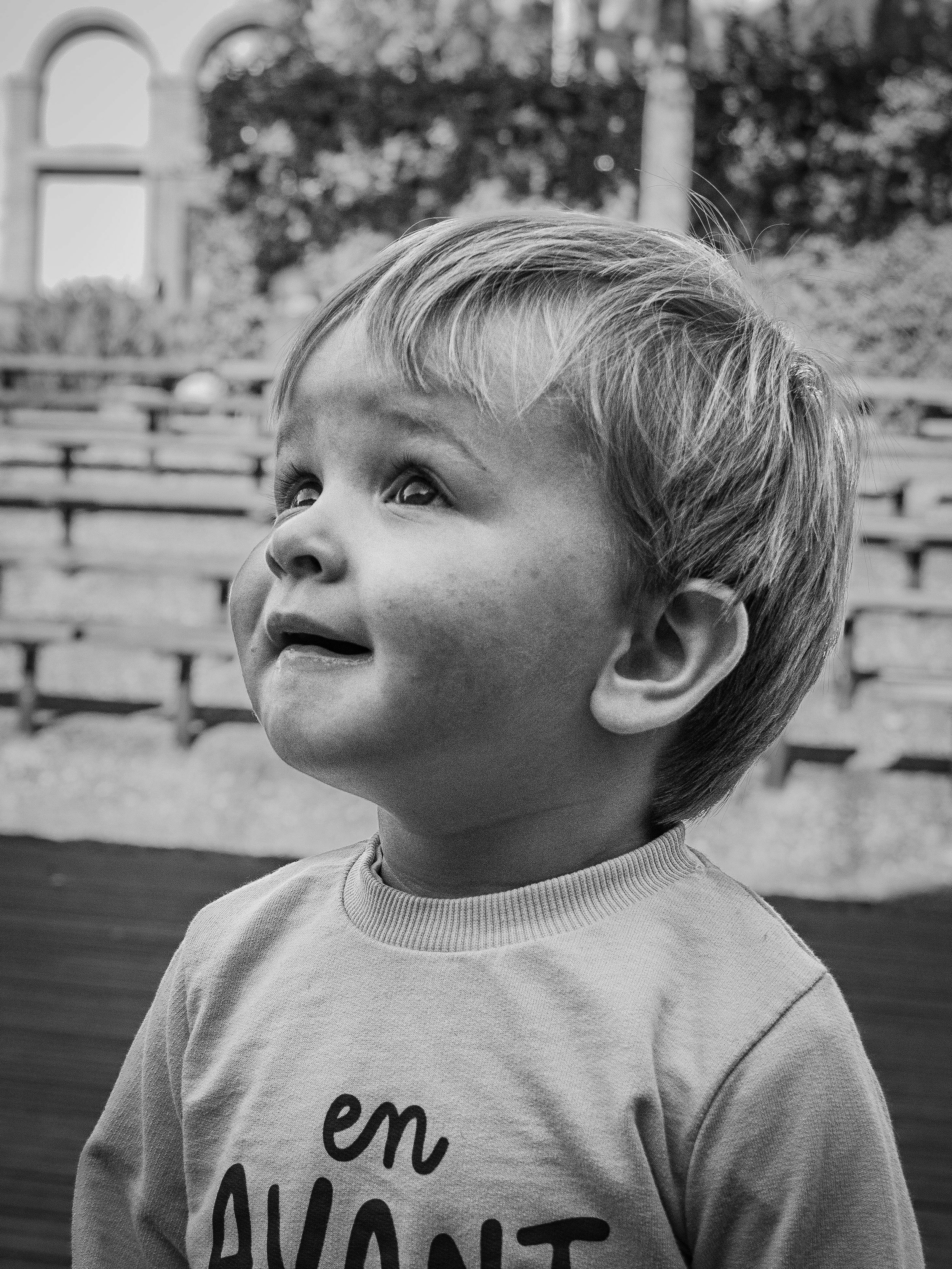 Un jeune garçon regarde vers le haut avec une expression de curiosité ou d'émerveillement, en noir et blanc, en plein air avec des gradins en bois en arrière-plan.
