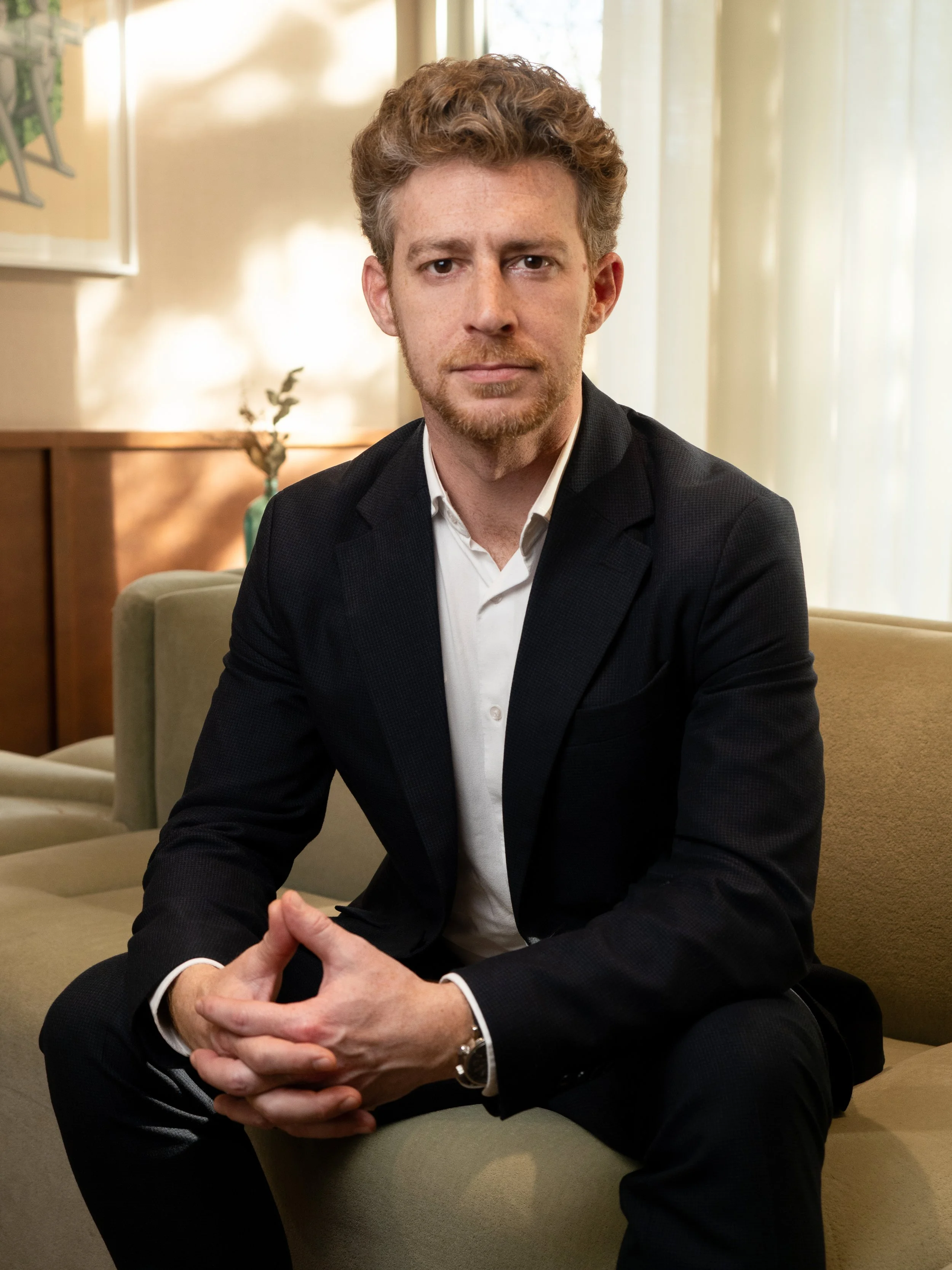 Un homme en costume noir assis sur un canapé beige, dans une pièce lumineuse avec rideaux clairs et une lampe, posture confiante avec les mains jointes.