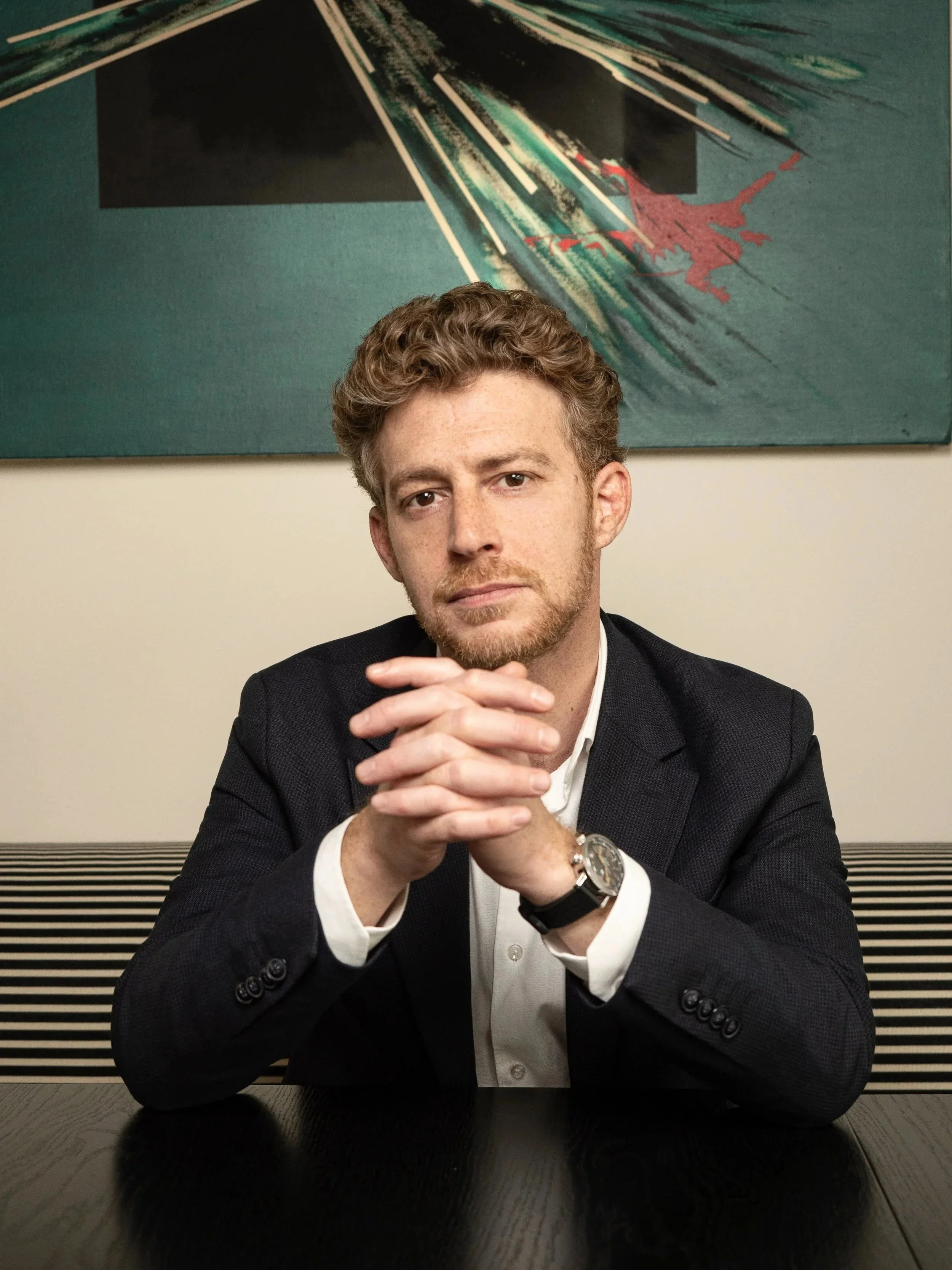 Jeune homme assis à une table en tenant ses mains jointes devant lui, portant une veste noire, une chemise blanche et une montre. Art abstrait en arrière-plan.
