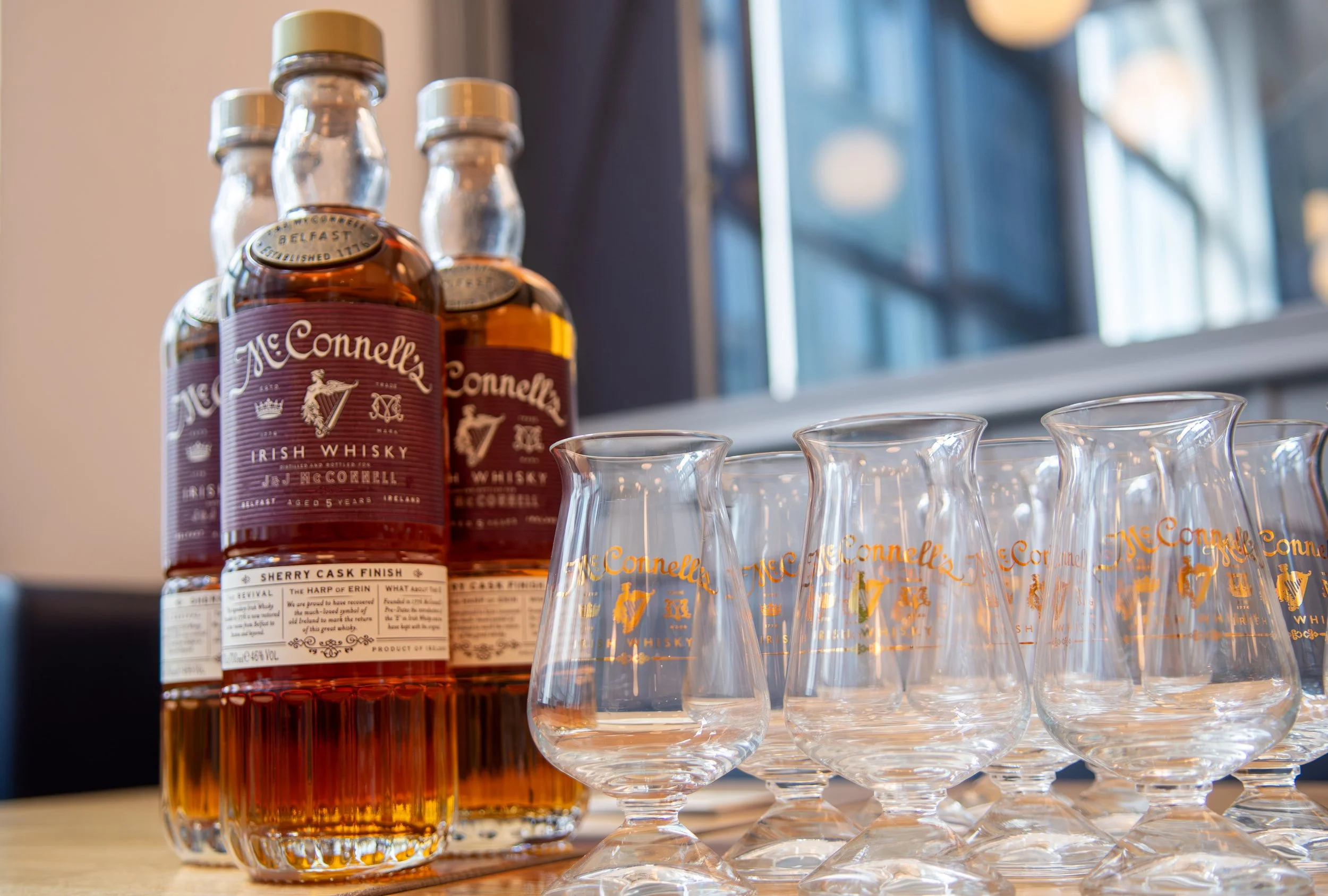 Three bottles of McConnell's Irish Whiskey and six whiskey glasses with orange and gold lettering, arranged on a wooden table near a window.