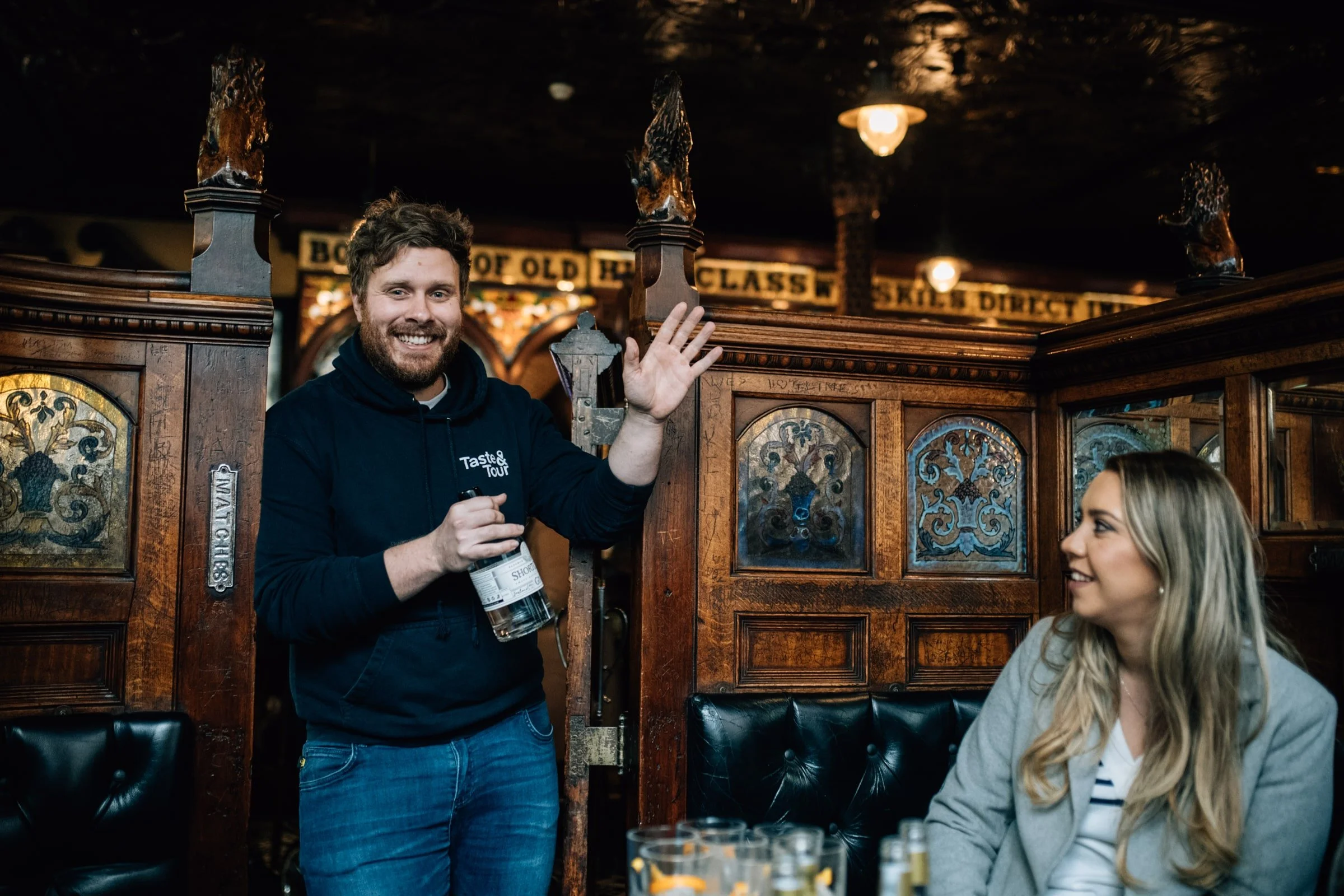 A man smiling and waving, holding a bottle of shot, standing next to a seated woman in a pub with wooden details and decorative stained glass panels.
