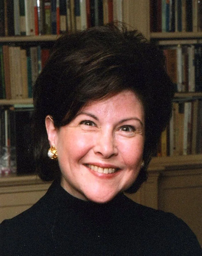 A woman with dark, styled hair, wearing a black top and gold earrings, smiling in front of a bookshelf filled with books.