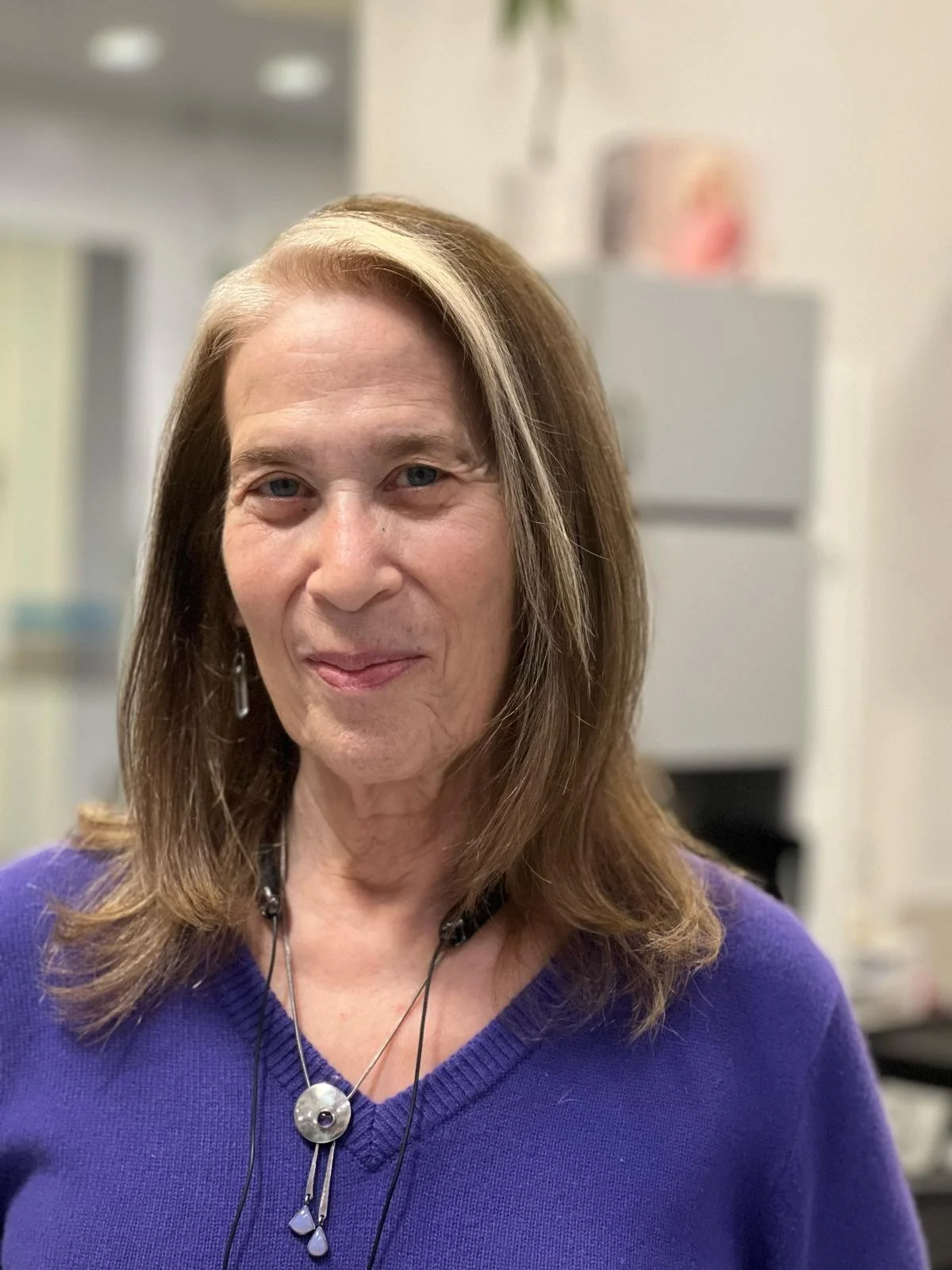 A middle-aged woman with shoulder-length brown hair and blonde highlights, wearing a purple sweater and jewelry, standing indoors with a blurred background of a room or office.