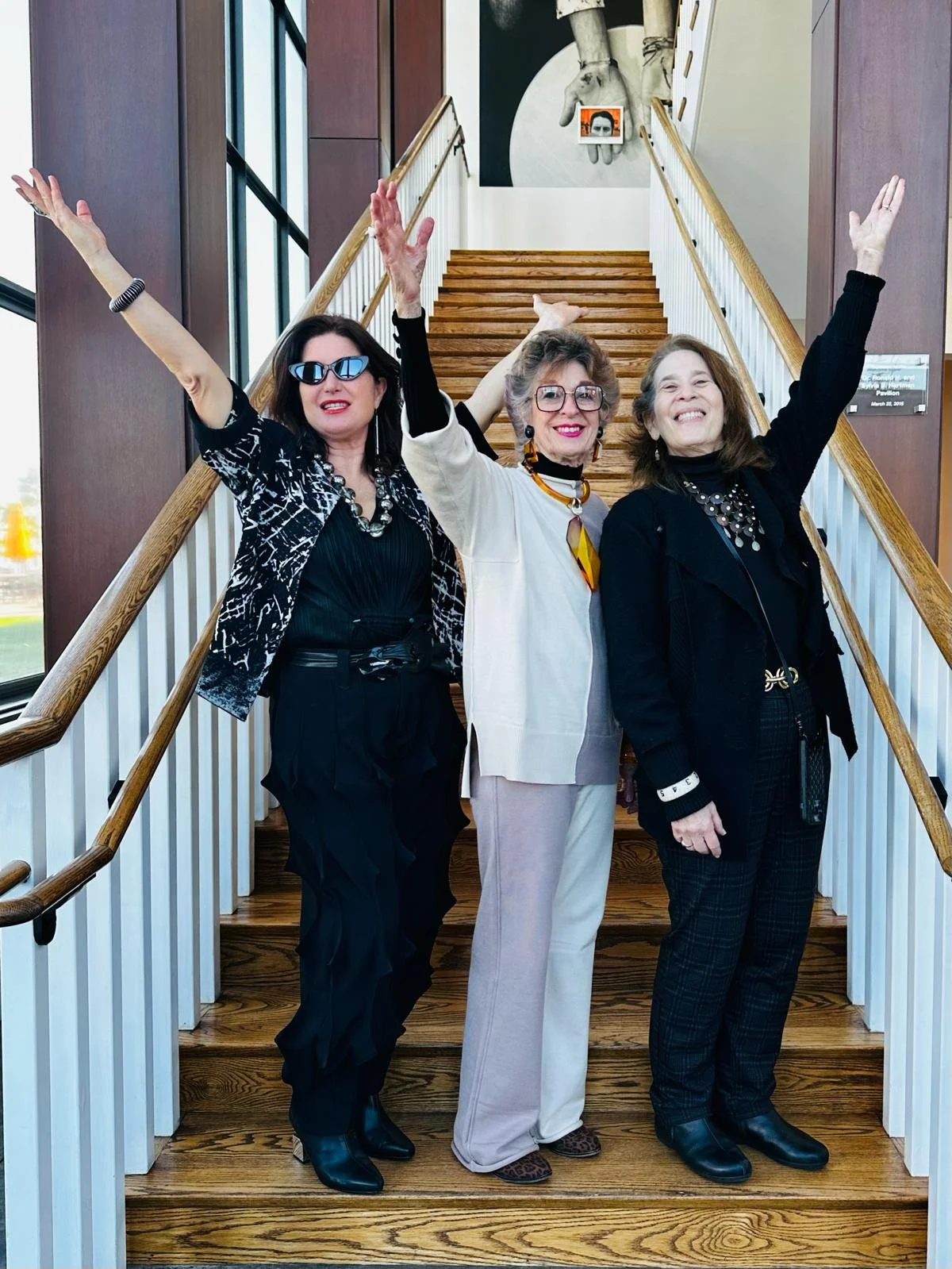Three women standing on a wooden staircase inside a building, smiling and raising their arms in celebration.