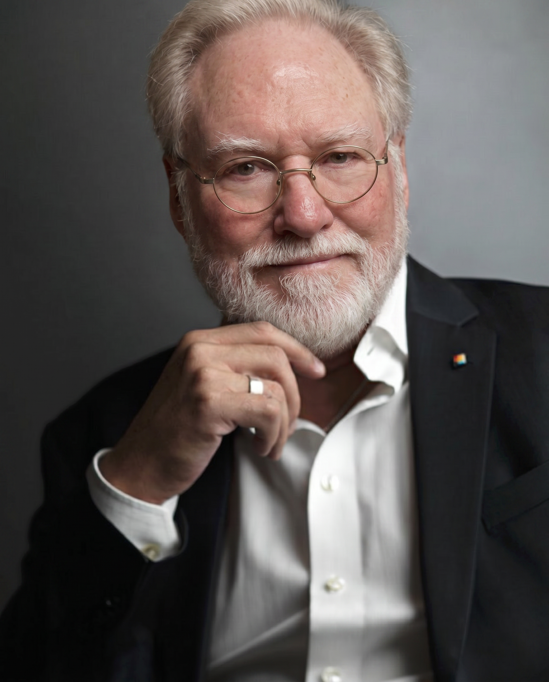 A man with a beard holding glasses, looking at the camera against a dark background.