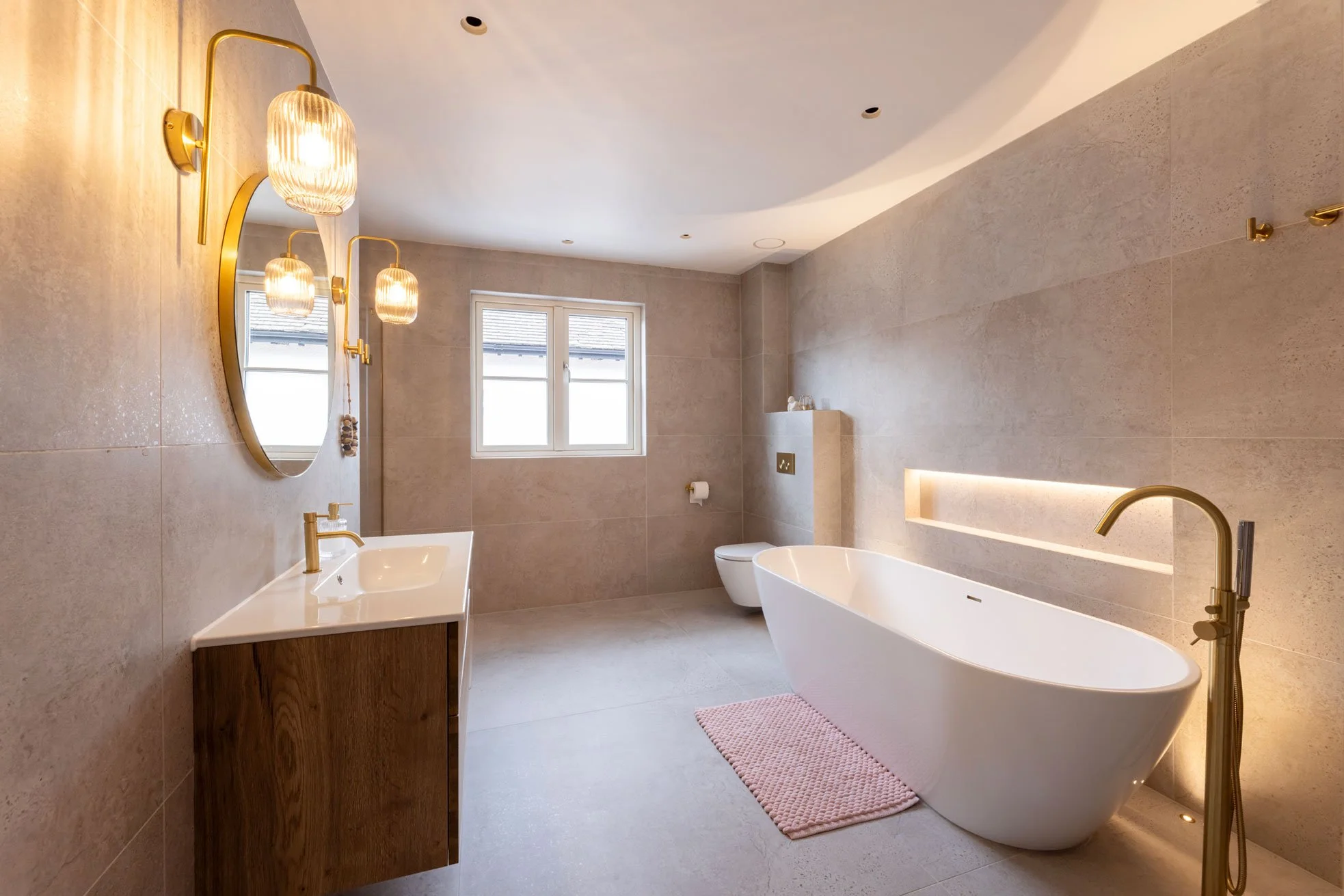 Modern bathroom with a freestanding bathtub, wooden vanity with a white sink, round mirror, wall-mounted lights, beige tiled walls and floor, window, and pink bath mat.