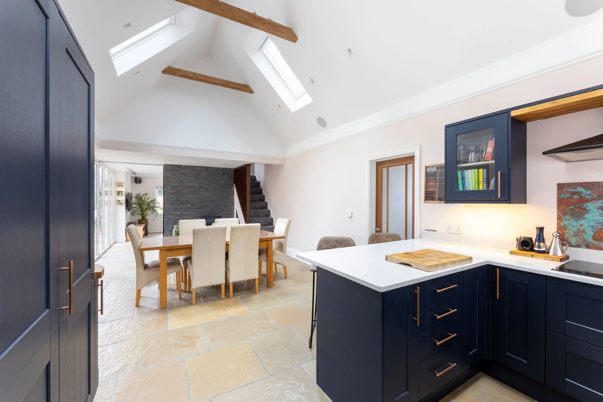 Modern open-plan kitchen and dining area with navy blue cabinets, white countertops, a wooden dining table with beige chairs, skylights, and a staircase with dark carpet leading upstairs.