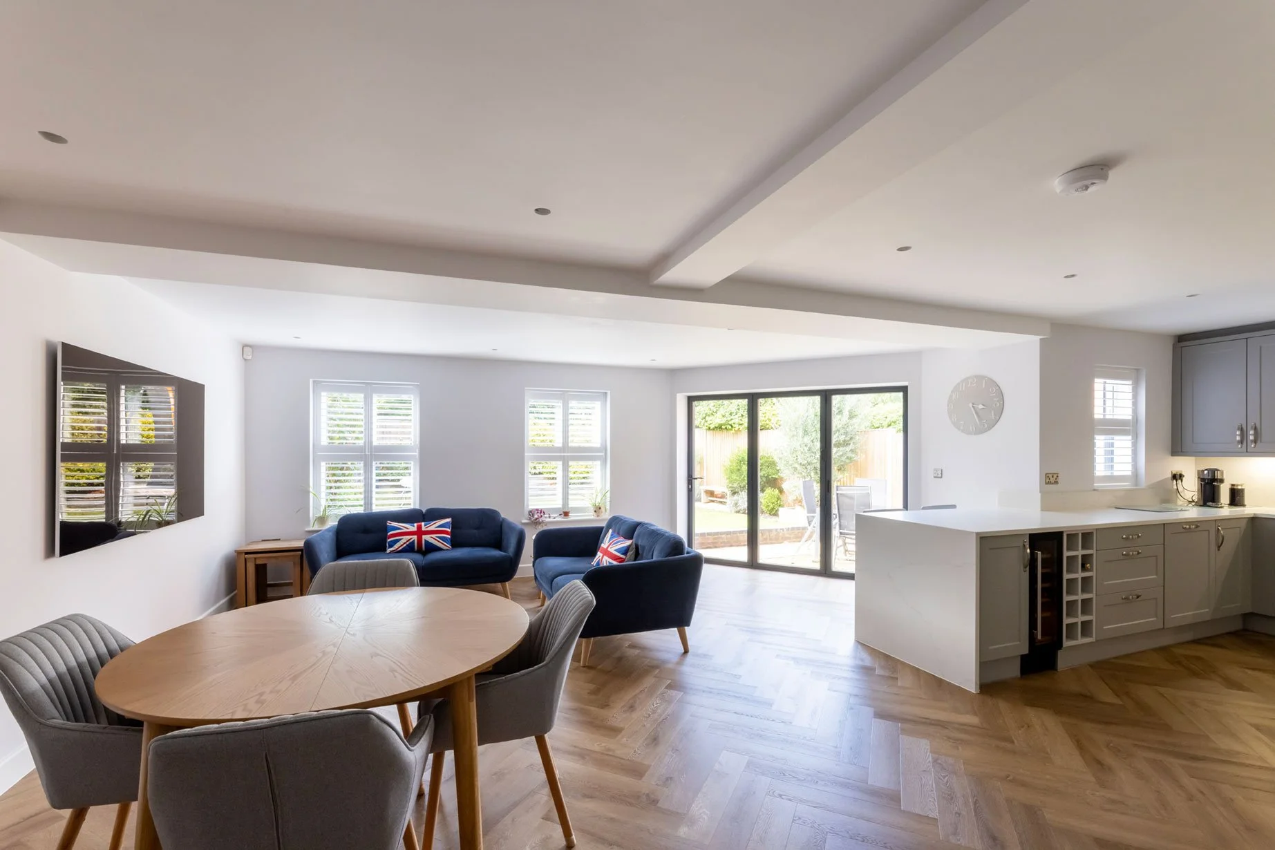 Open-concept living room and kitchen with large windows, sliding glass door to backyard, wooden flooring, gray cabinets, wall clock, wall-mounted TV, and seating with British flag pillows.