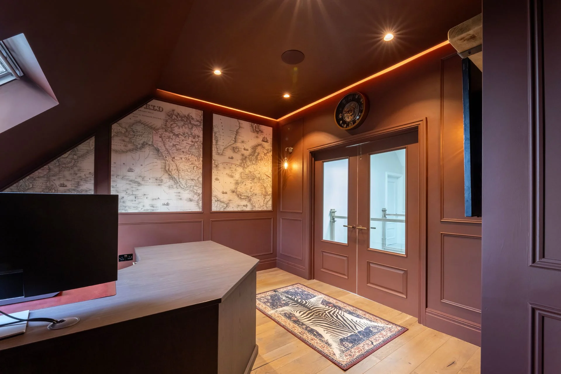 A room with pinkish walls, a wooden floor, and a rug with zebra and floral patterns, featuring double glass doors. Wall art includes two large vintage maps and a decorative clock. There is a desk with a computer monitor and some electrical outlets, and a window with a small window ledge.