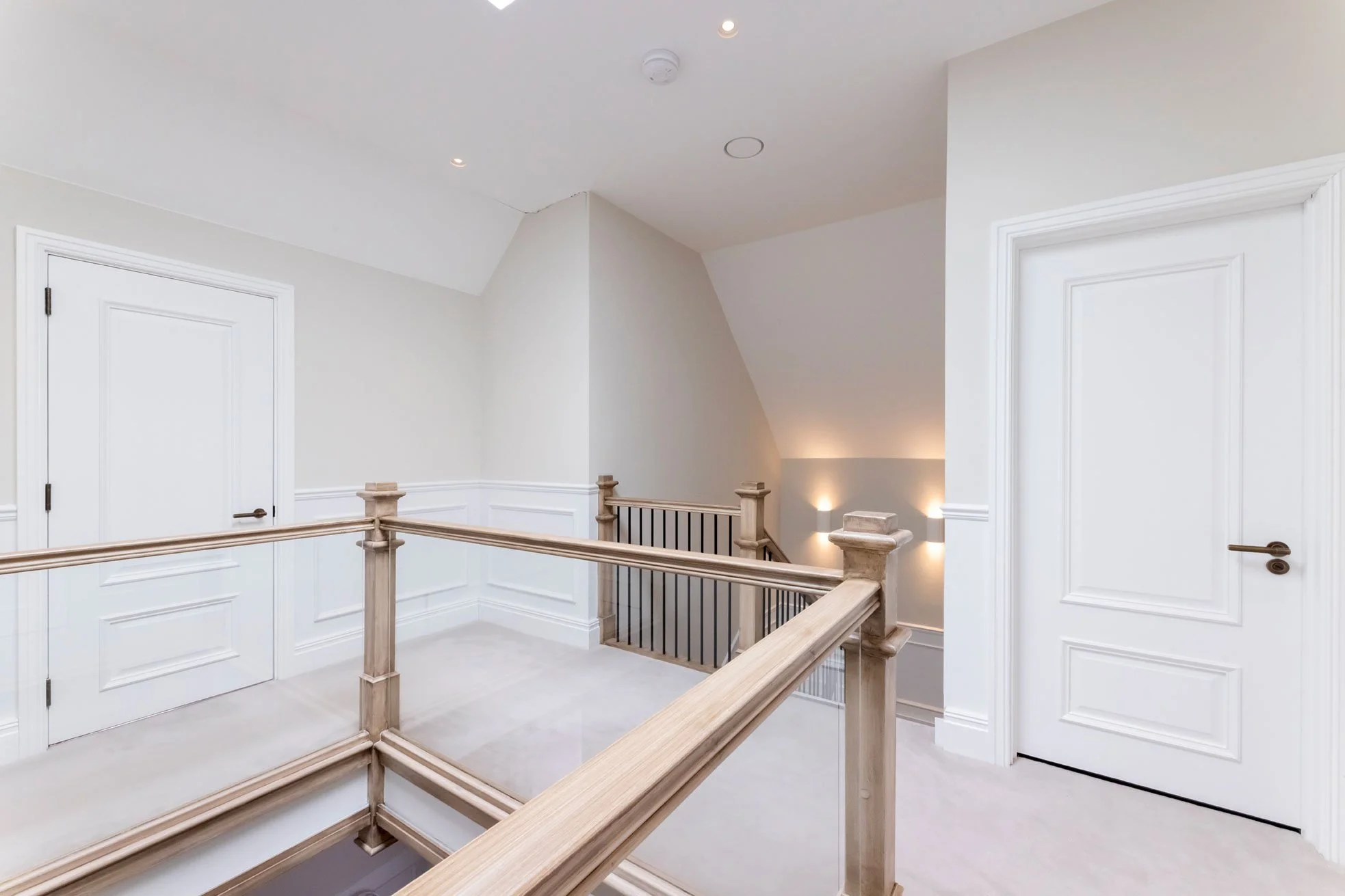 Empty upstairs landing area with white walls, two closed white doors, and a wooden and black metal staircase railing at the edge of the staircase, with wall-mounted lights illuminating the space.
