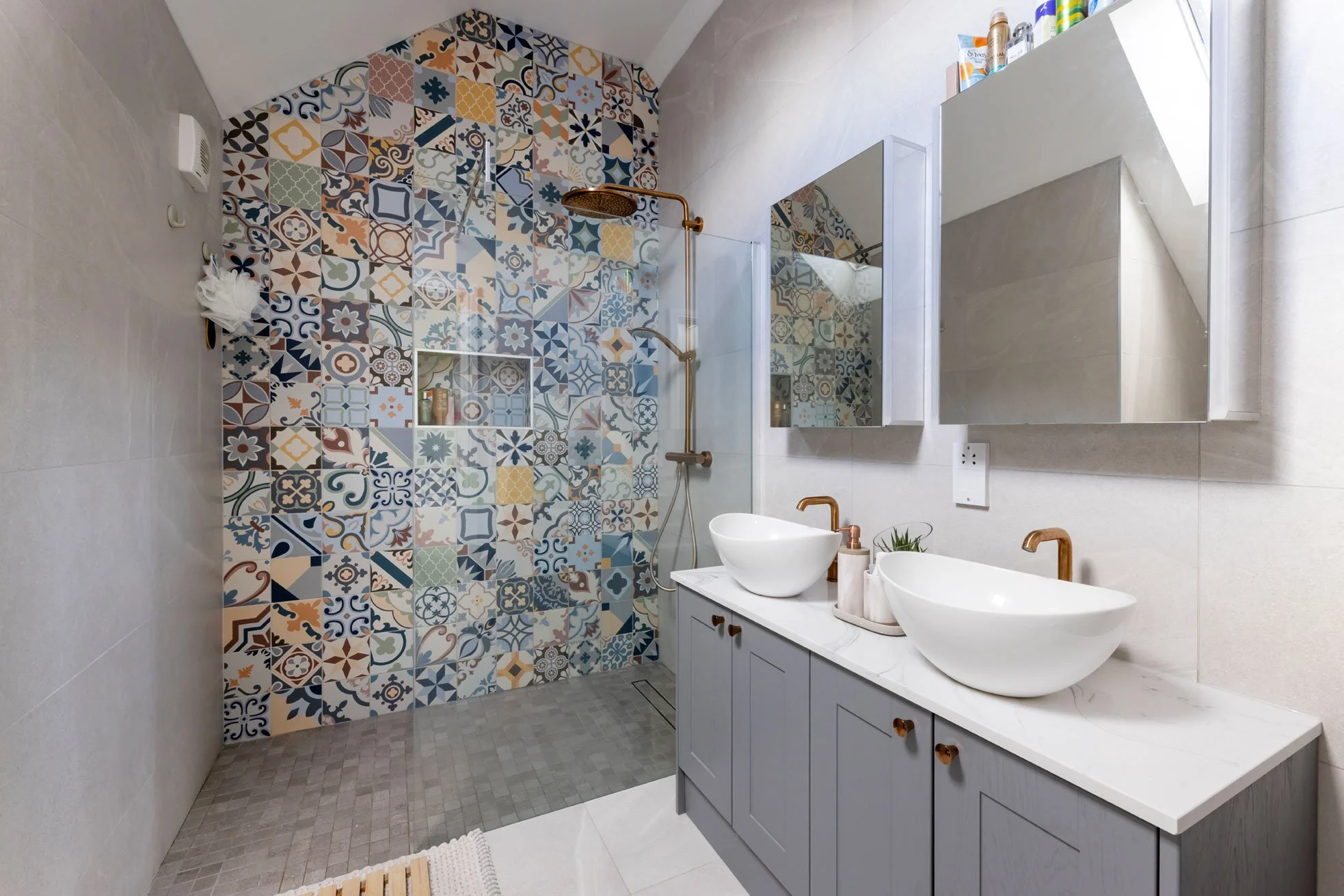 Modern bathroom featuring a colorful patterned tiled shower wall, double vessel sinks on a marble countertop, and light gray cabinets with copper hardware. There are two mirrors above the sinks, and items on the right storage cabinet, with natural light coming through a window.