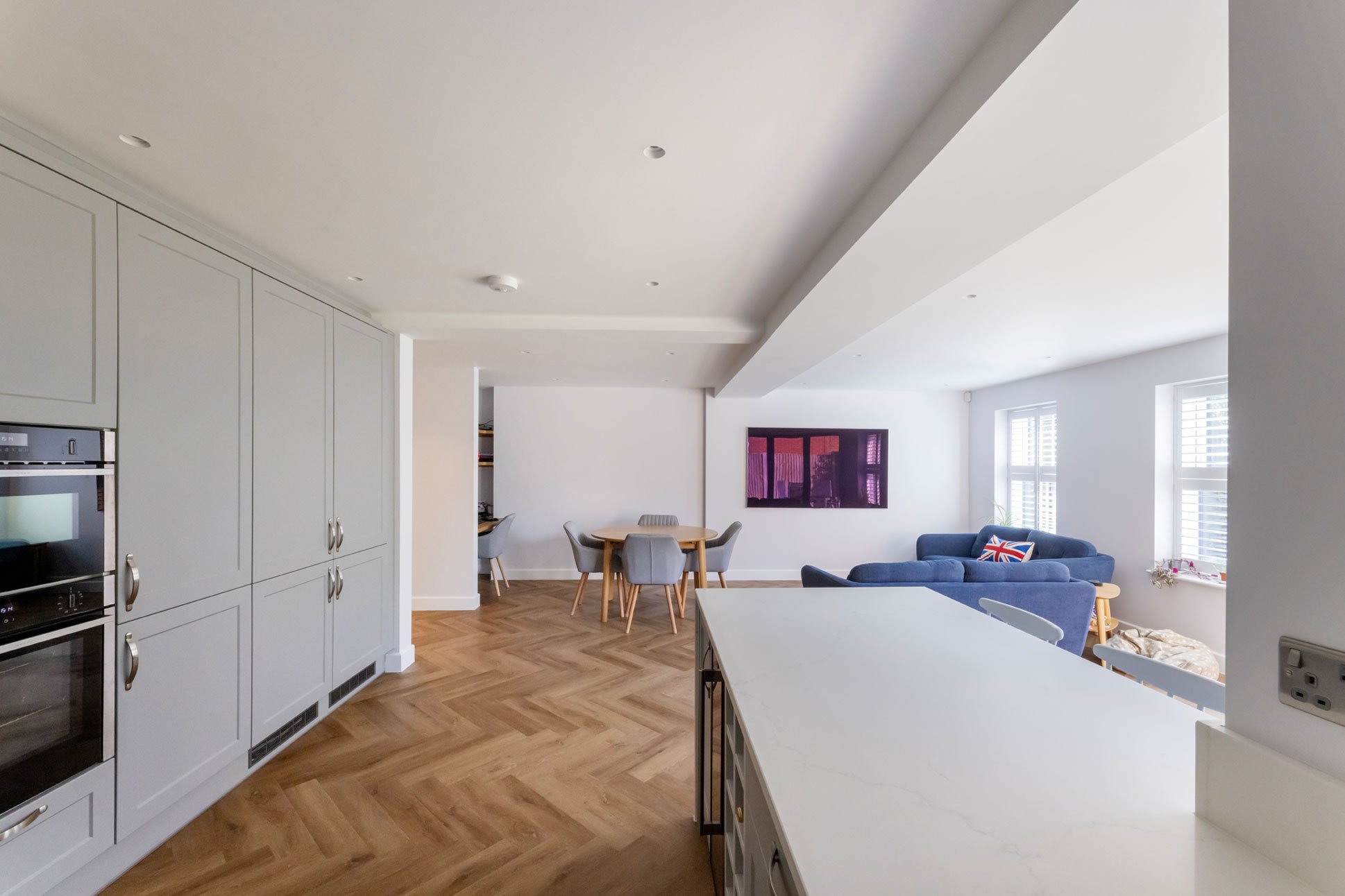 Open-plan living area with white kitchen cabinets, a dining table with gray chairs, a blue sofa with a Union Jack cushion, and large windows with white shutters.