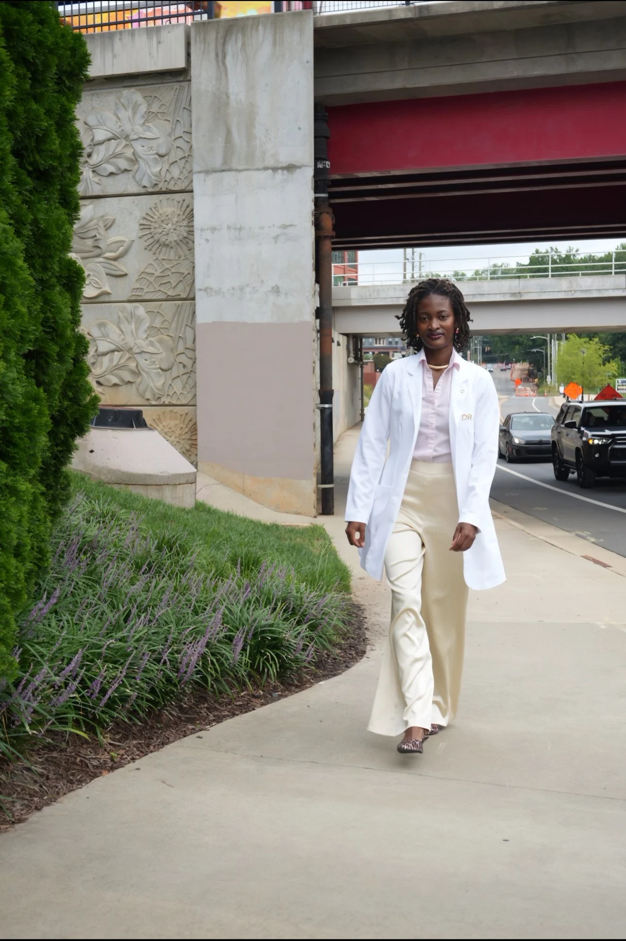 A woman in a white lab coat walking on a sidewalk under a bridge, with cars on the street behind her and decorative stone carvings on a wall to her left.