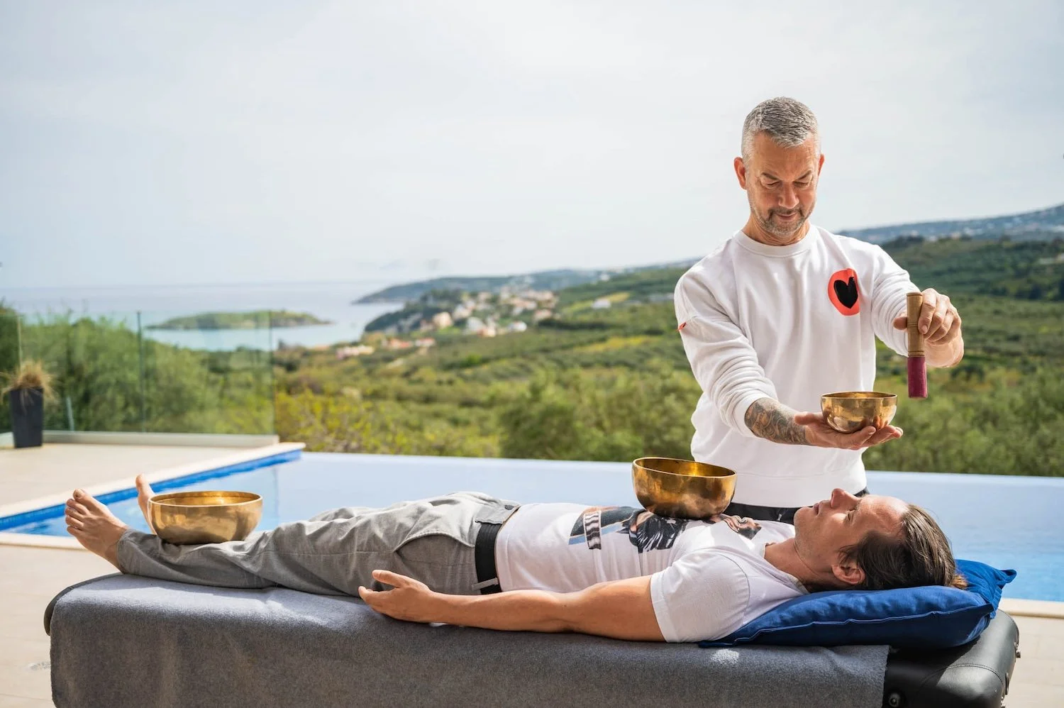 A man receiving sound therapy with Tibetan bowls outdoors near a pool, overlooking a scenic landscape of greenery and water, while lying on a massage table.