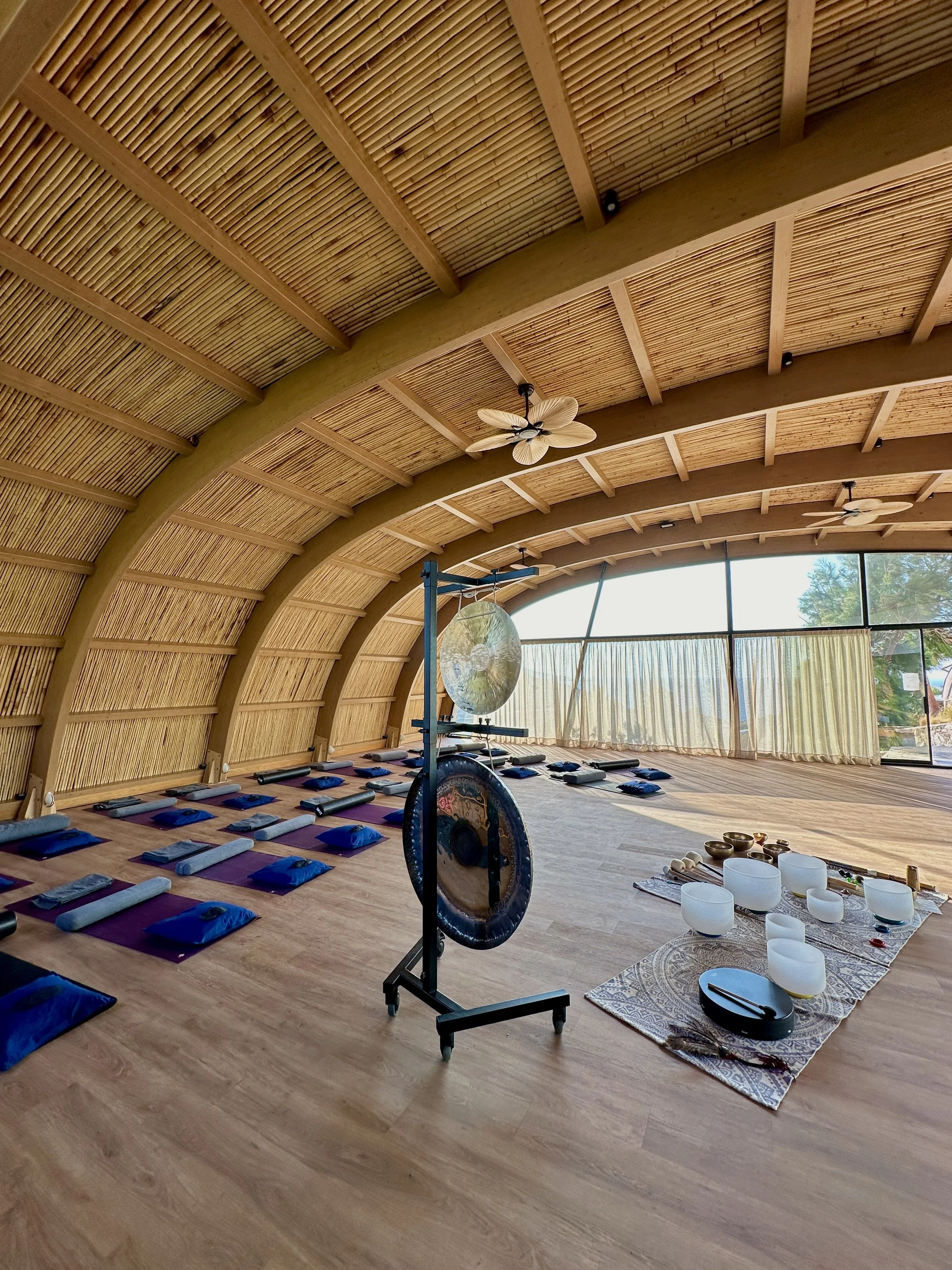 Interior of a spacious yoga or meditation studio with wooden arched ceiling, mats and bolster cushions on the floor, singing bowls and a gong on a stand, some wind chimes, and large windows with curtains.