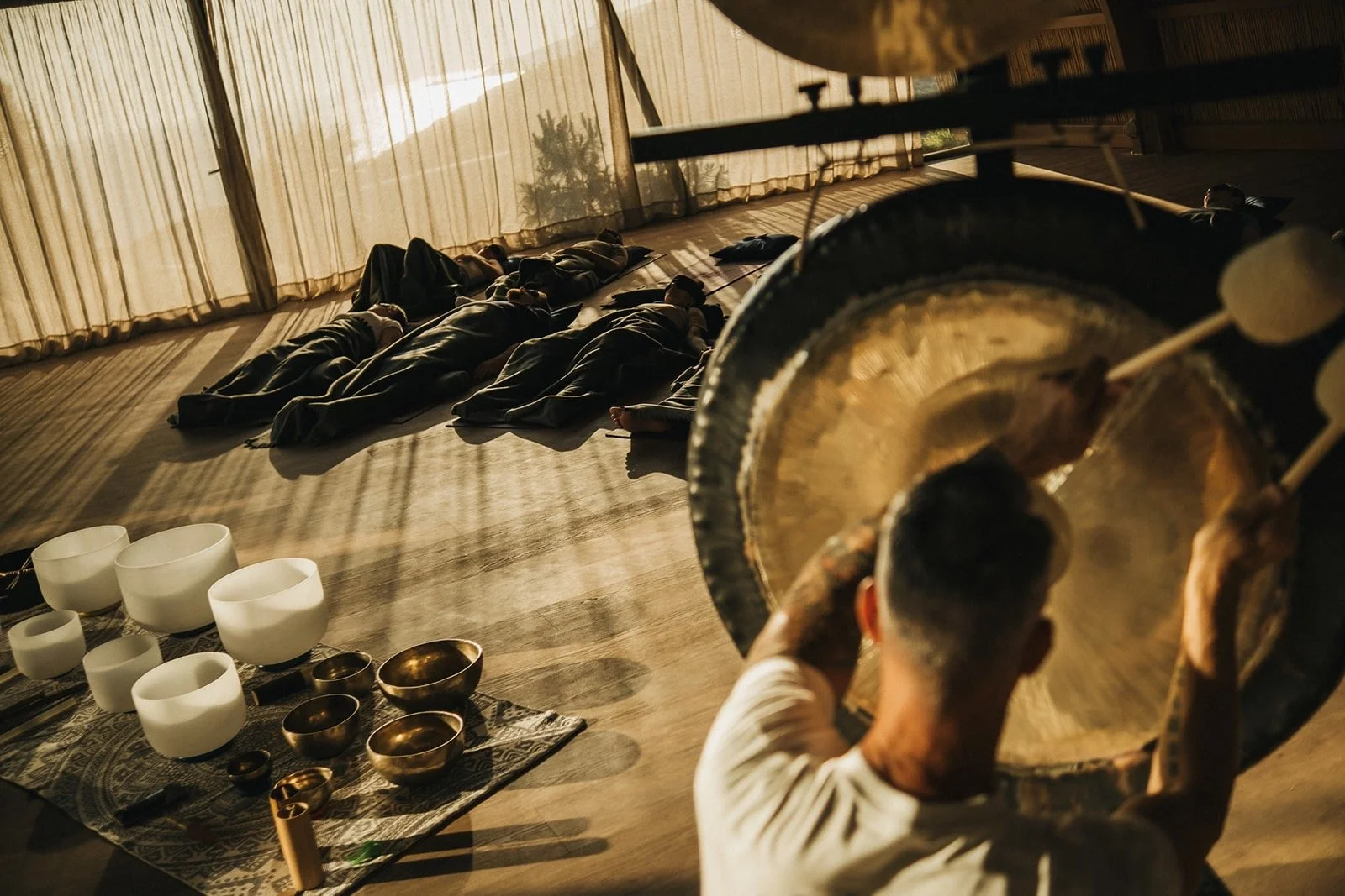People lying on the floor during a sound bath session, with a person playing a large gong and singing bowls arranged nearby, in a room with large windows and curtain.