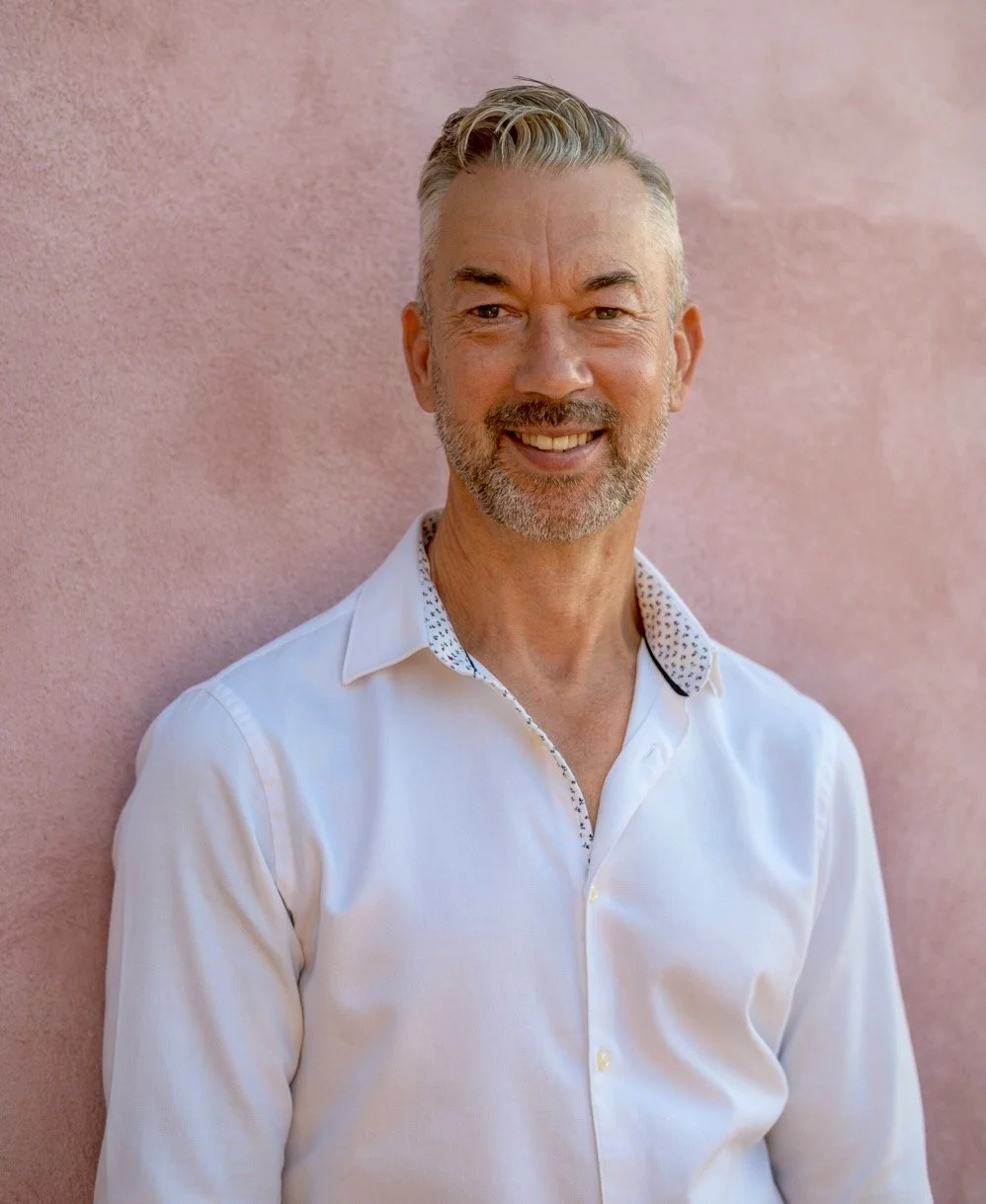 A smiling middle-aged man with gray hair and a beard, wearing a white shirt, standing against a pink textured wall.