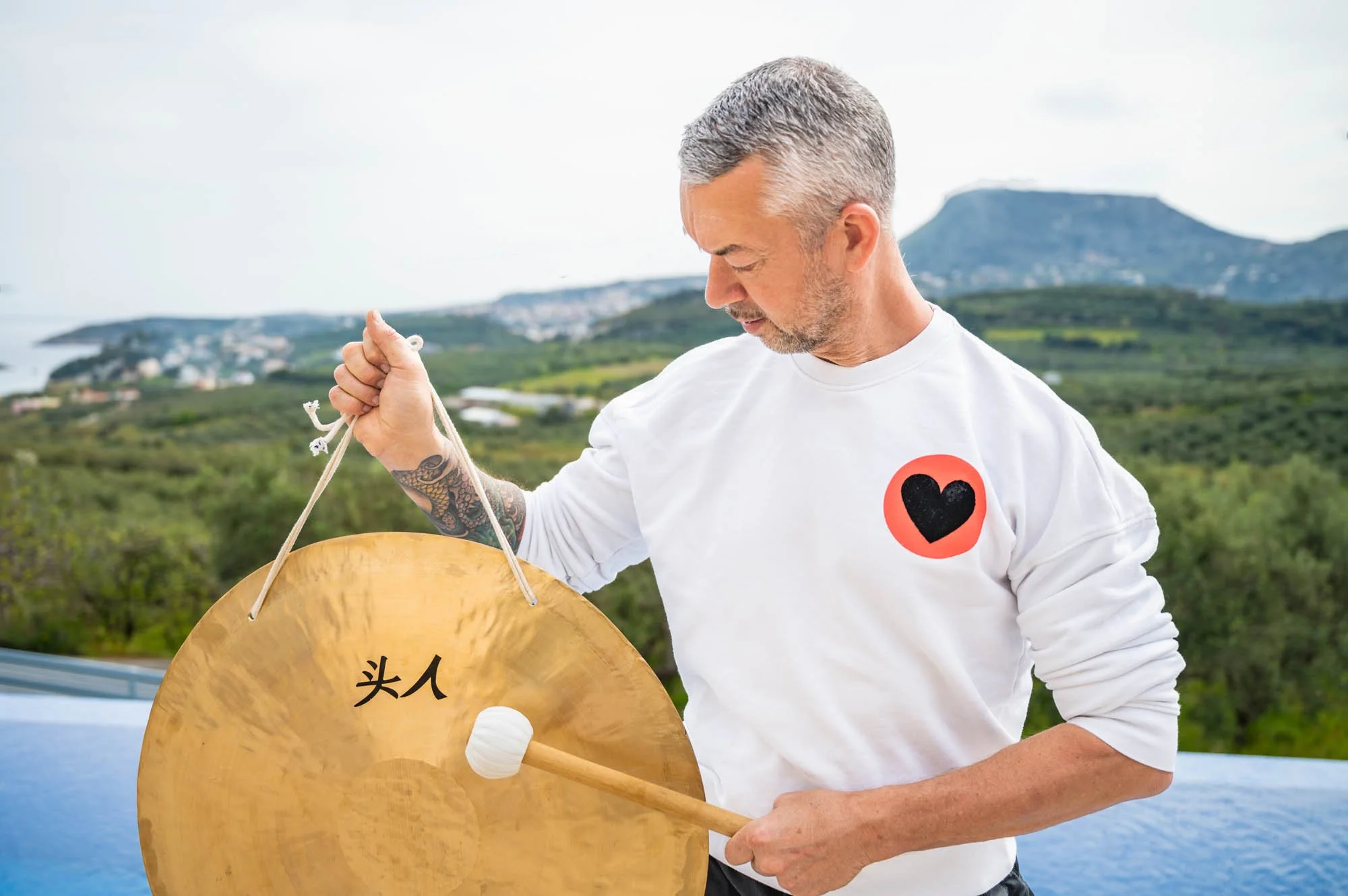 rob sykes sound therapist wearing a white sweatshirt  holding a chinese wind gong and a mallet, outdoors with a scenic landscape of greenery, mountains, and water in the background.