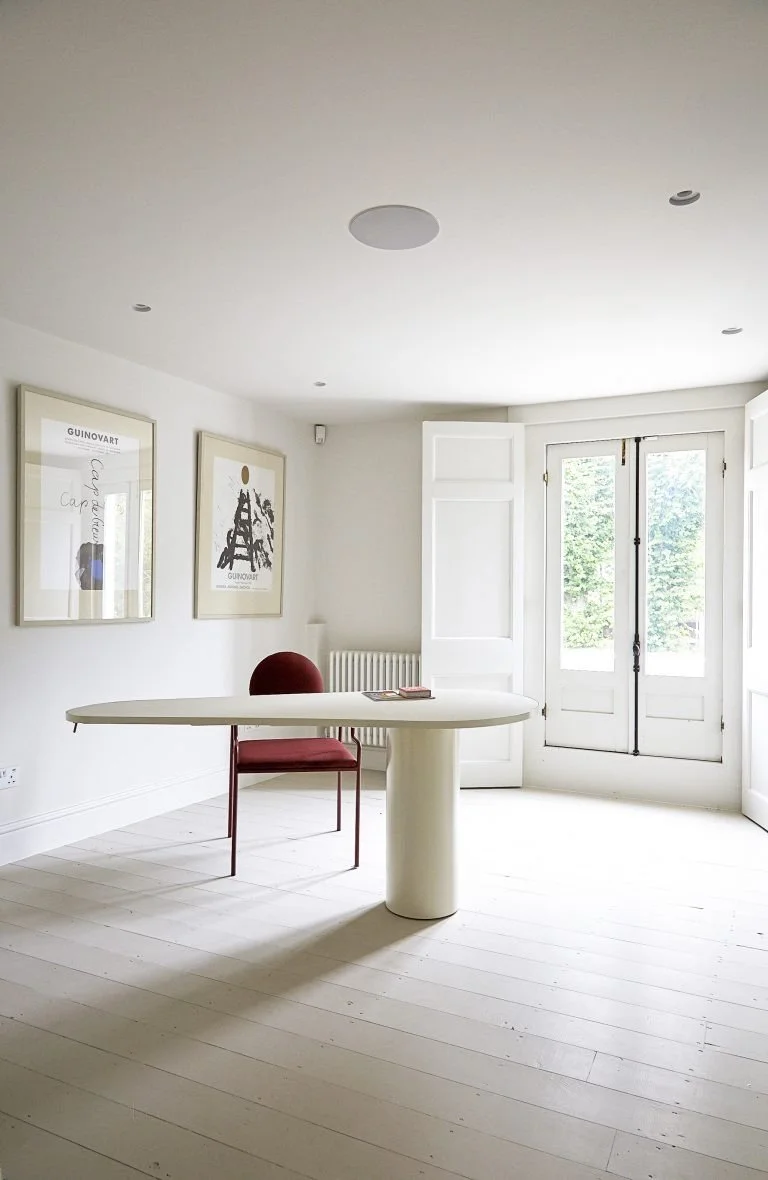Bright room with white walls and floor, a white modern table with rounded edges, a red chair, framed artwork on the wall, and tall white doors opening to a view of greenery outside.