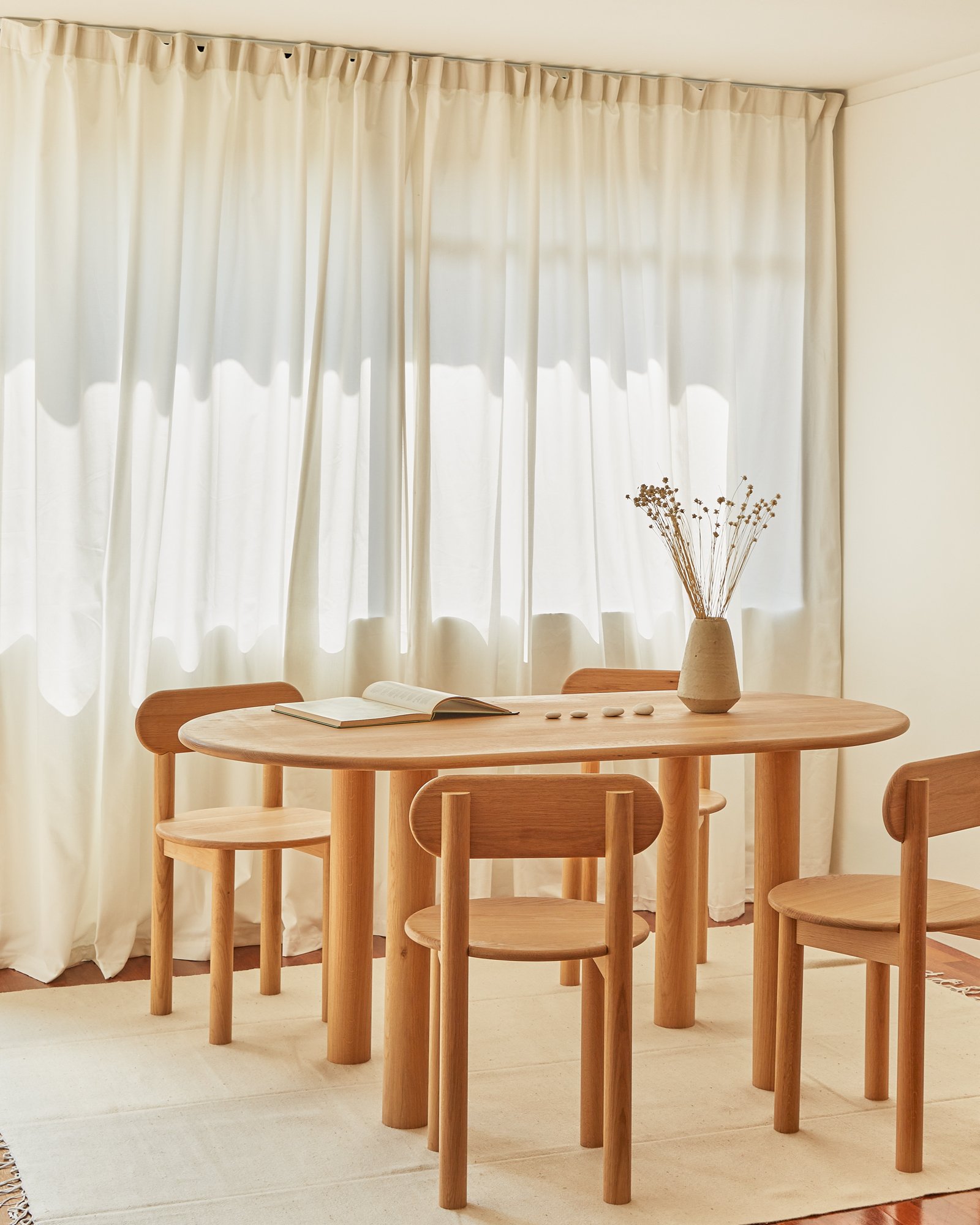 A wooden dining table with four matching chairs in front of a window with white curtains. The table has an open book, a beige vase with dried flowers, and small stones on it. The room has neutral-colored walls and a light-colored rug.