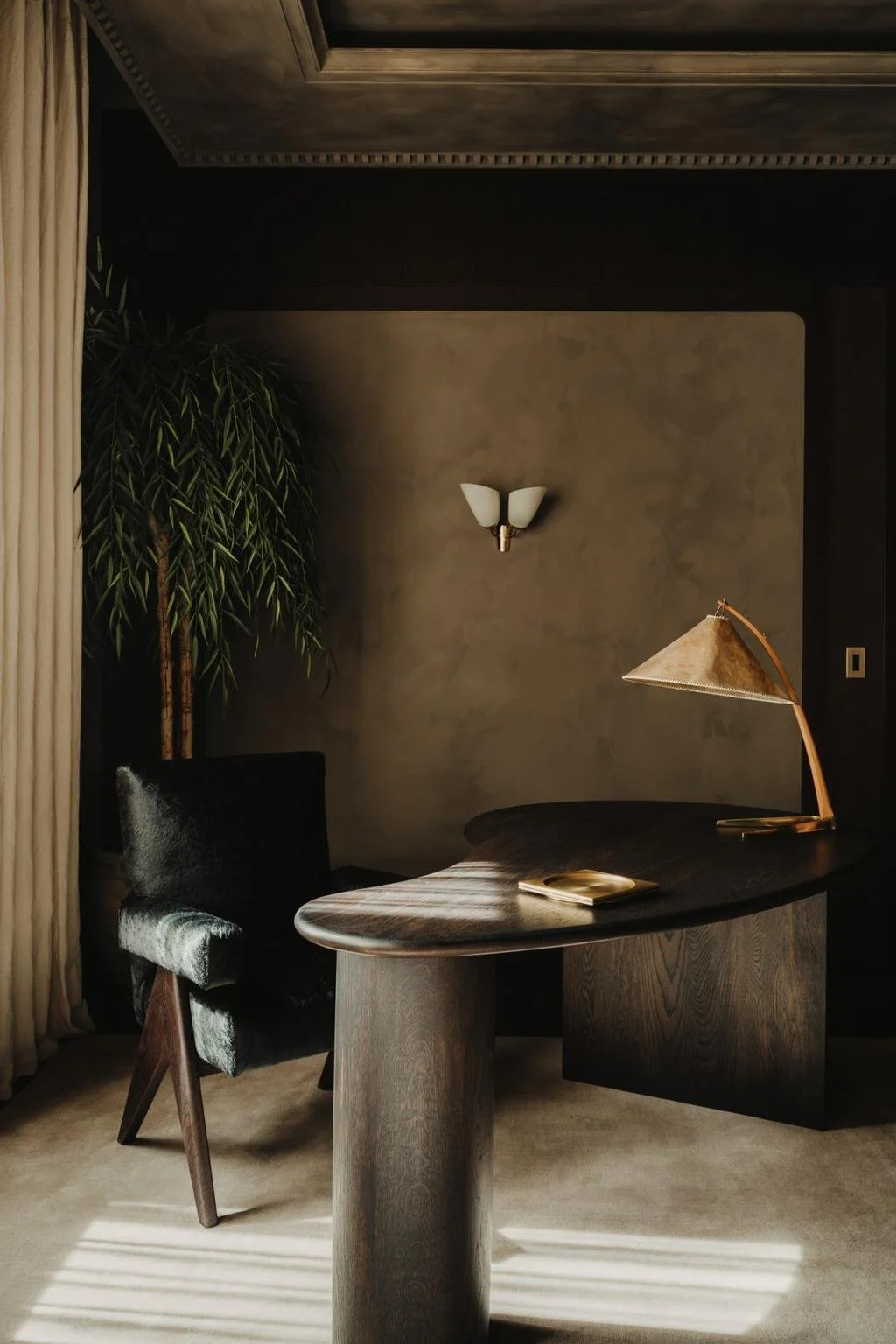A dimly lit office with a dark wood desk, a black velvet chair, a gold desk lamp, a decorative wall sconce, and a potted plant in the corner.