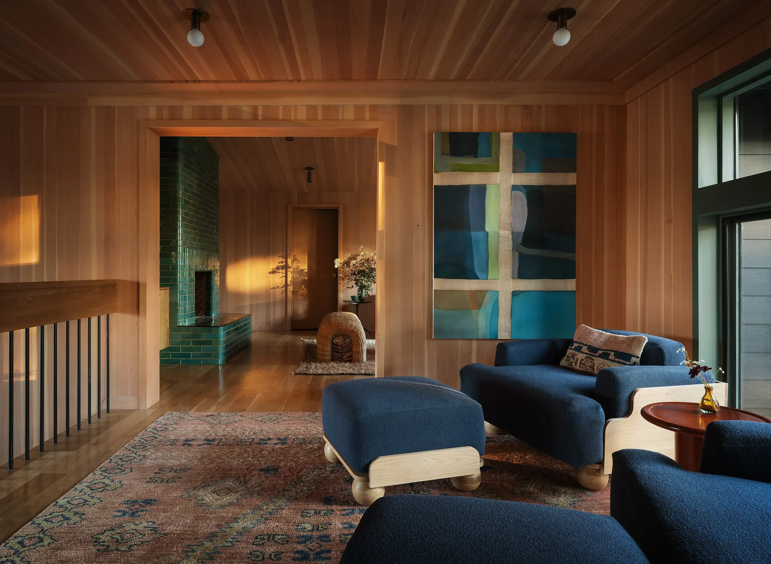 A cozy living room with wood-paneled walls and ceiling, blue furniture, a patterned rug, and modern artwork on the wall. Sunlight streams in through large windows, casting warm light on the decor.