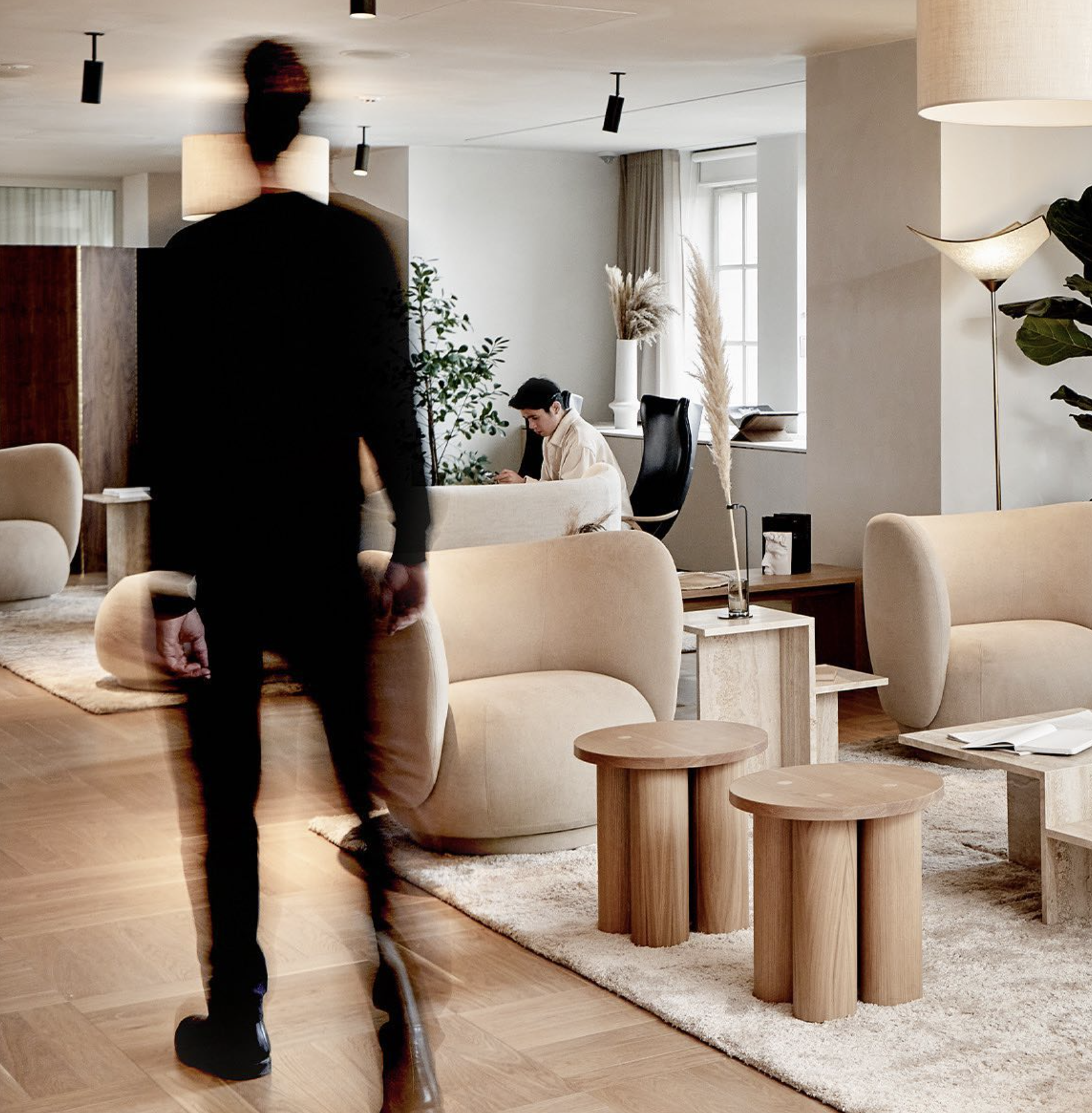 A modern, stylish living room with beige sofas, wooden stools, and a plush rug. Two people, one blurred in motion walking away and one sitting at a desk, are present in the room.
