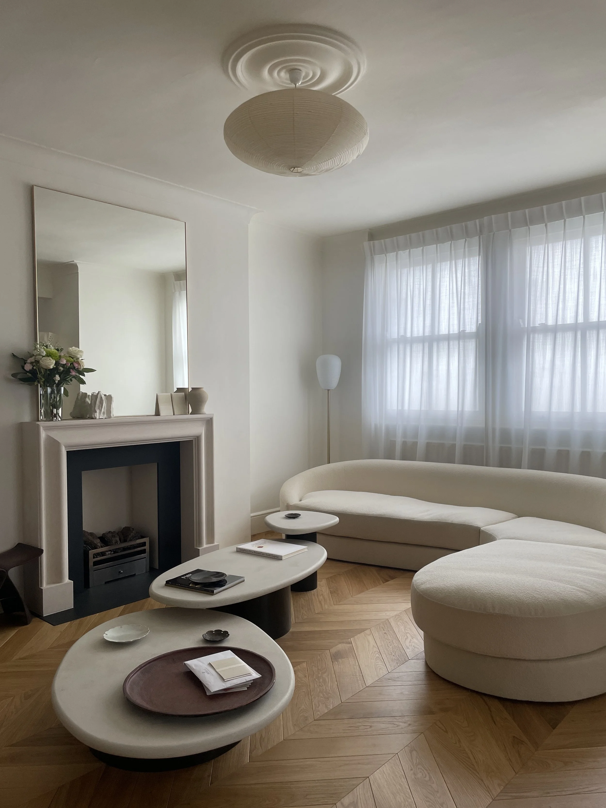Minimalist living room with white walls, a curved beige sofa, a fireplace with a mirror above it, a tall floor lamp, and multiple small coffee tables on a wooden parquet floor.