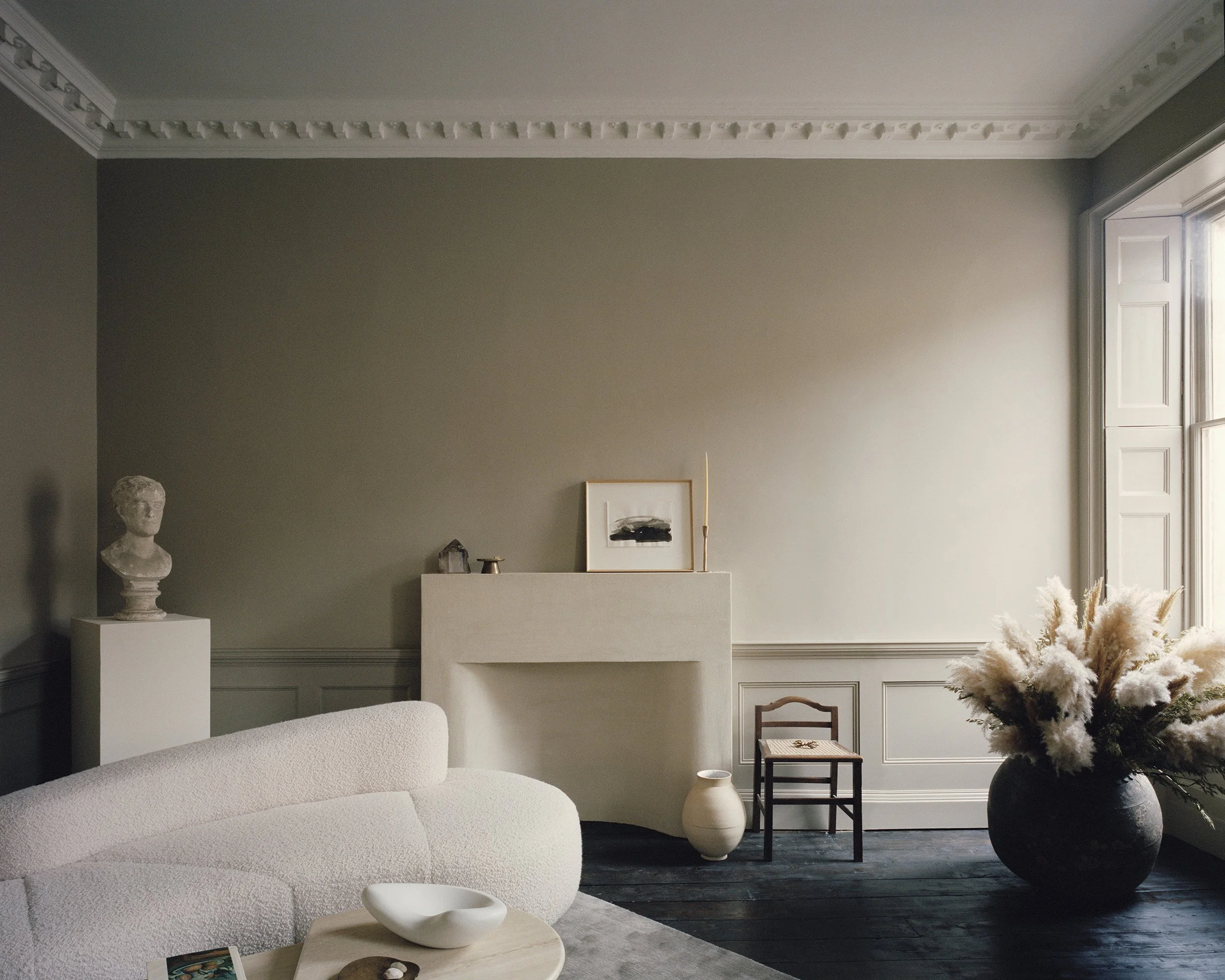 A minimalist living room with a beige wall, a white couch, a small table, a bust sculpture on a pedestal, and a large window with natural light, accented with pampas grass in a black vase.