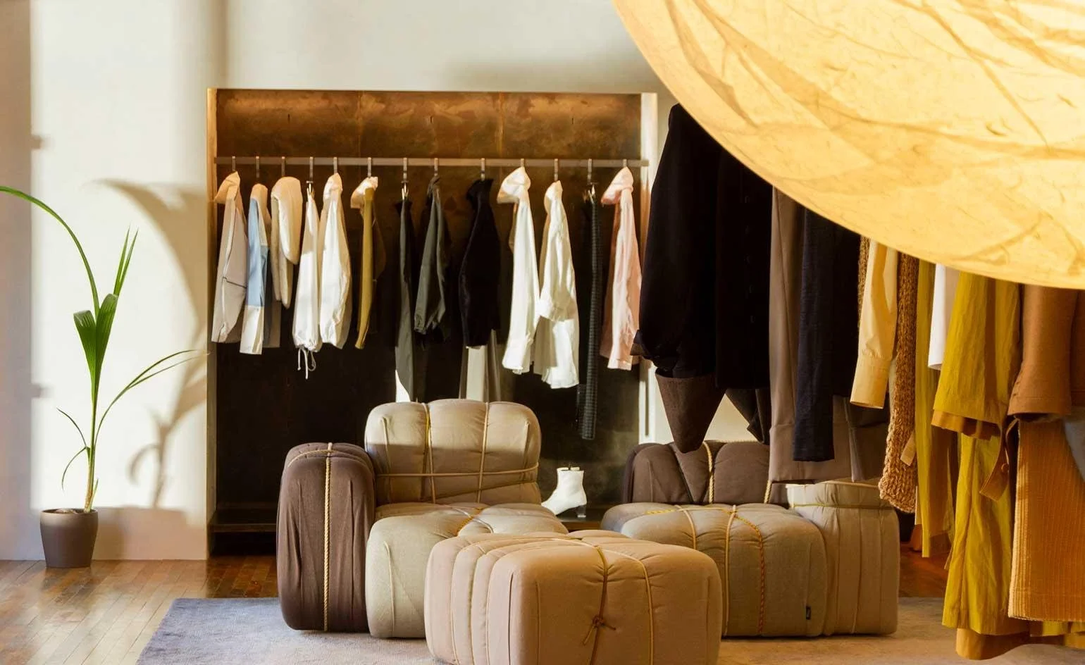 A cozy dressing room with hanging clothes, a beige armchair, matching ottoman, a small table with a lamp, a potted plant, and a section of yellow and brown clothing.