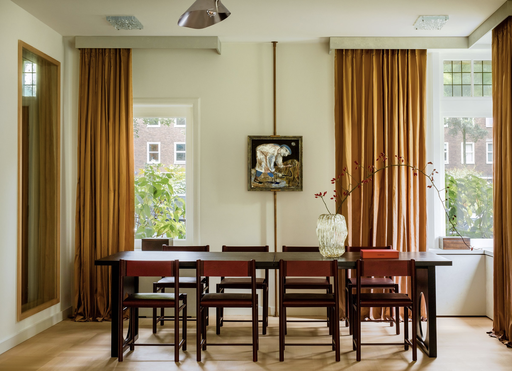 A dining room with a black table, six red chairs, a large glass vase with branches, yellow curtains, and windows showing an outdoor view of trees and brick buildings.