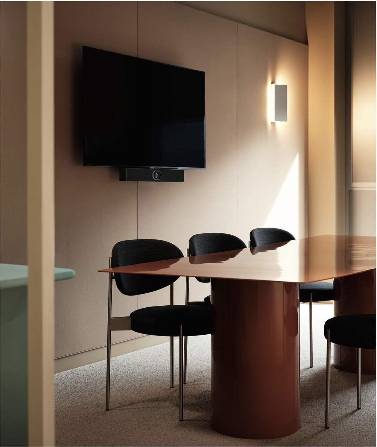 Meeting room with a wall-mounted flat-screen TV, a security camera below, a rectangular wooden table, and four black chairs, illuminated by sunlight and wall lights.