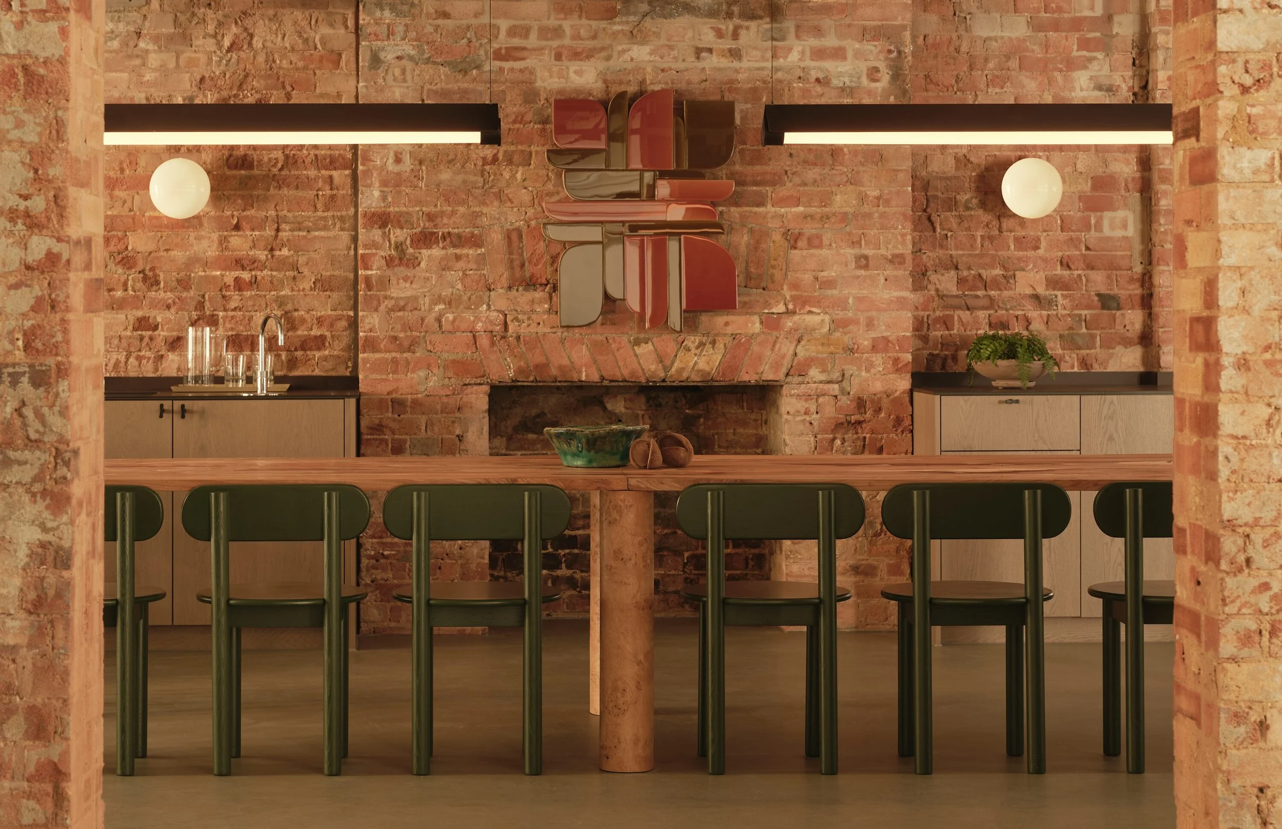 Interior of a cozy, modern dining area with a brick wall, a long wooden table surrounded by green chairs, minimal decor, and warm lighting.